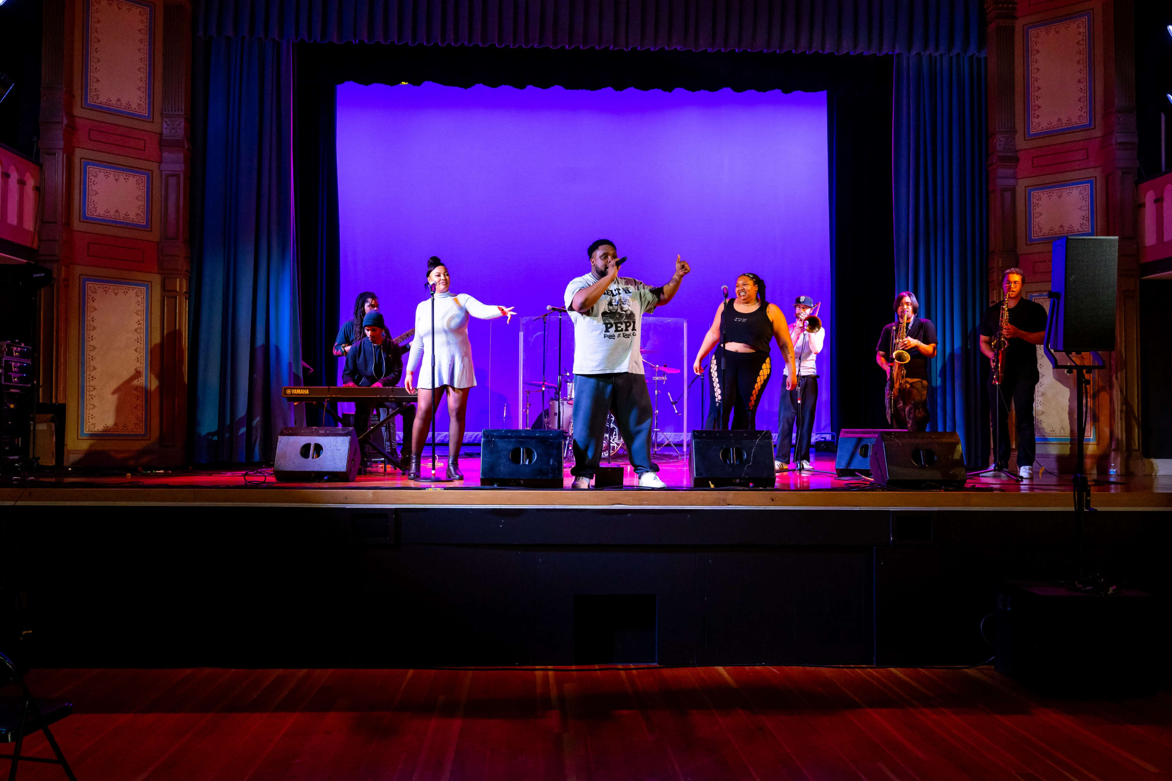 A diverse group of musicians performs on stage in a theater with blue and purple lighting.