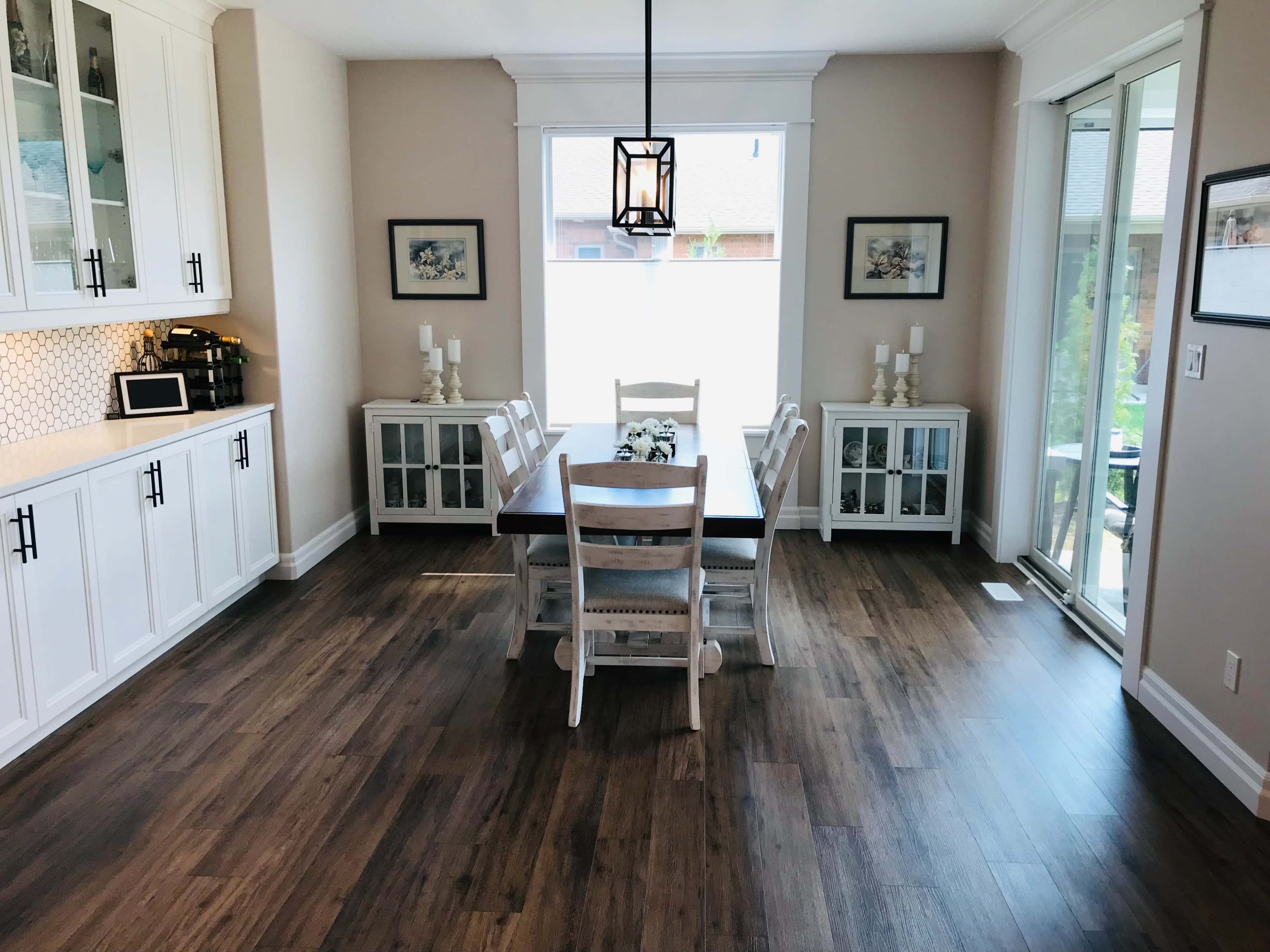 A dining room features a rectangular table surrounded by white chairs, with a light fixture above and cabinets with glass doors against the wall.