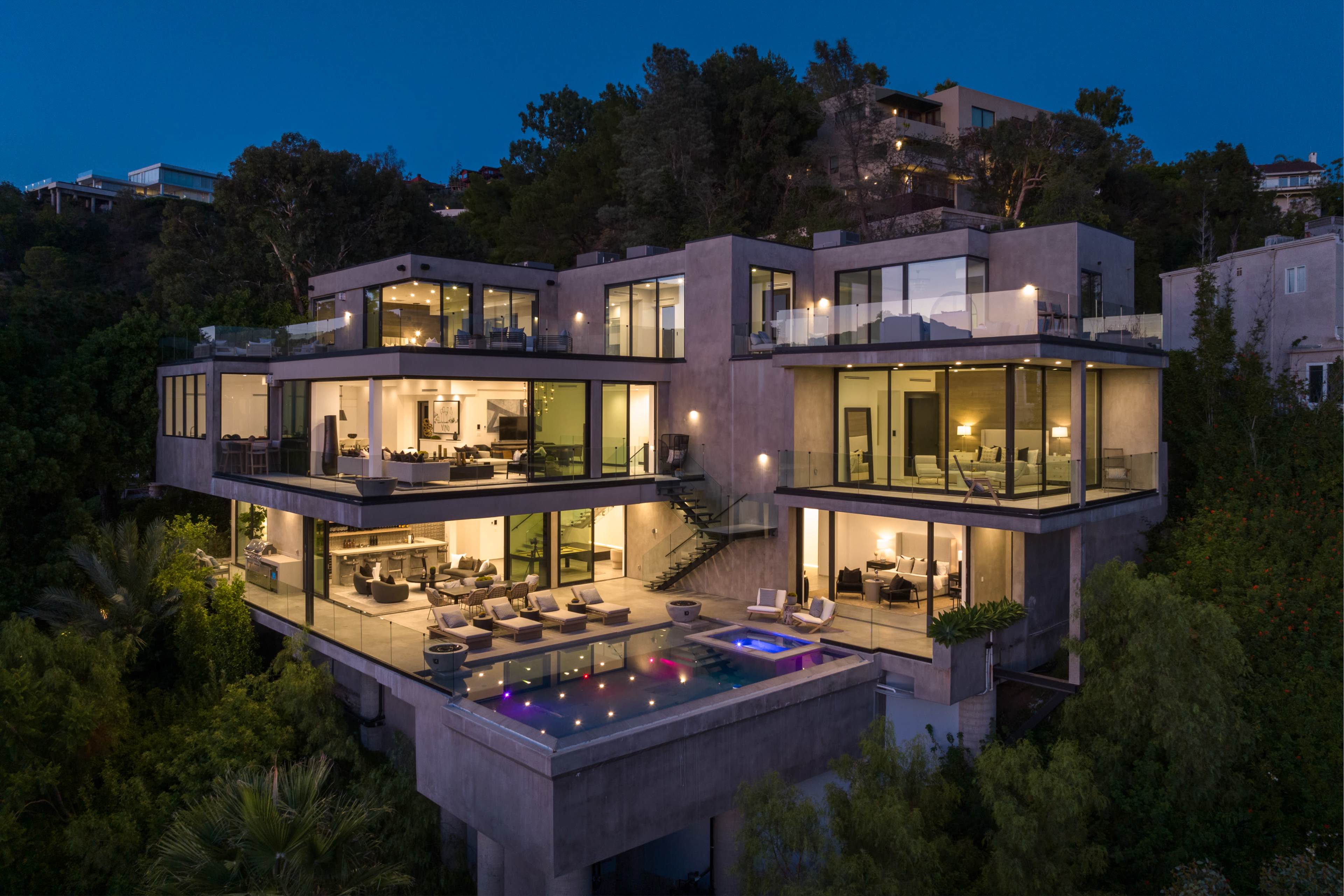 A modern multi-level house with large windows overlooks a pool area, surrounded by lush greenery and illuminated at night.