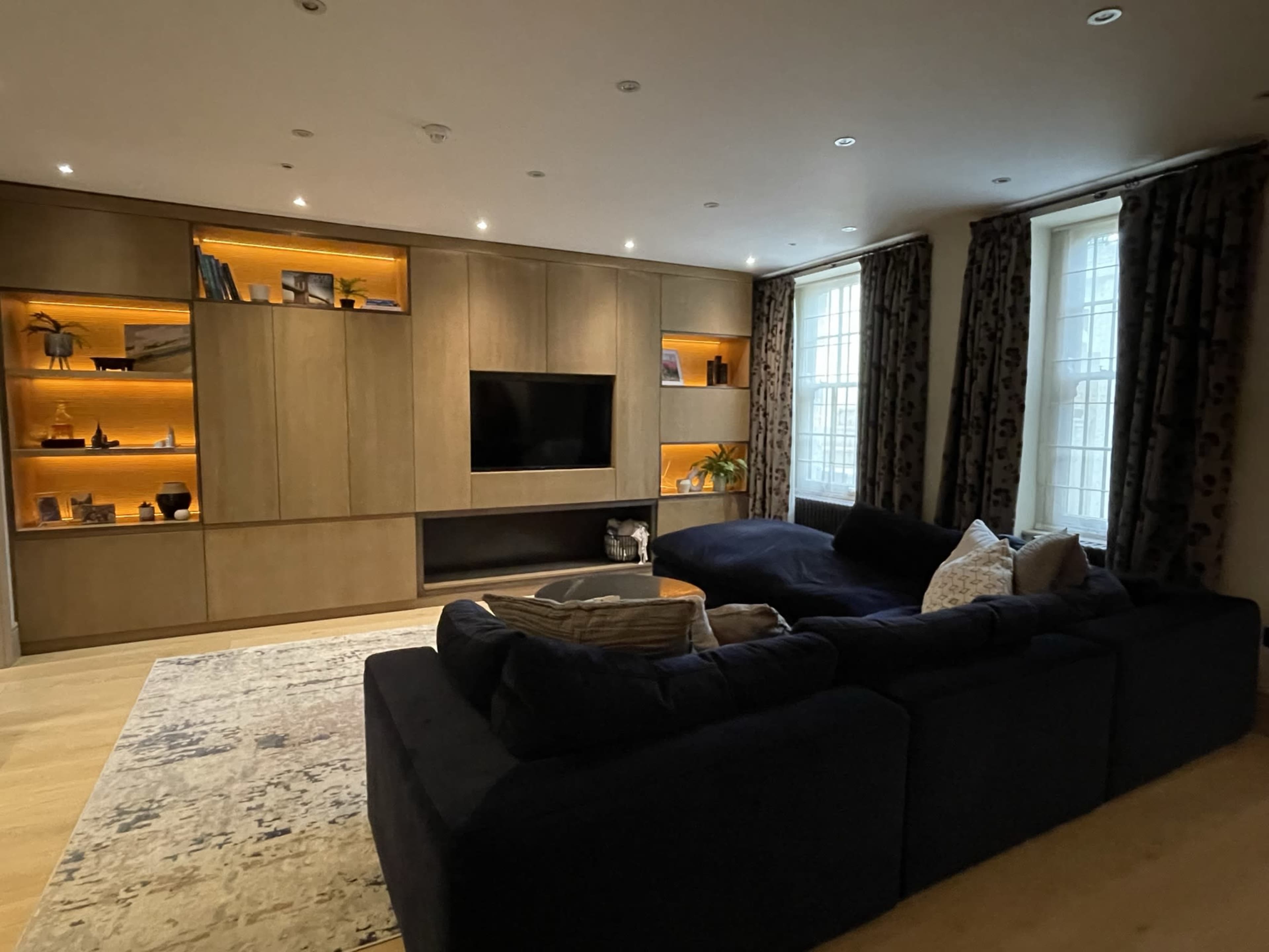 A modern living room with a dark sectional sofa, a large wall-mounted TV, and built-in shelves illuminated by warm lighting.