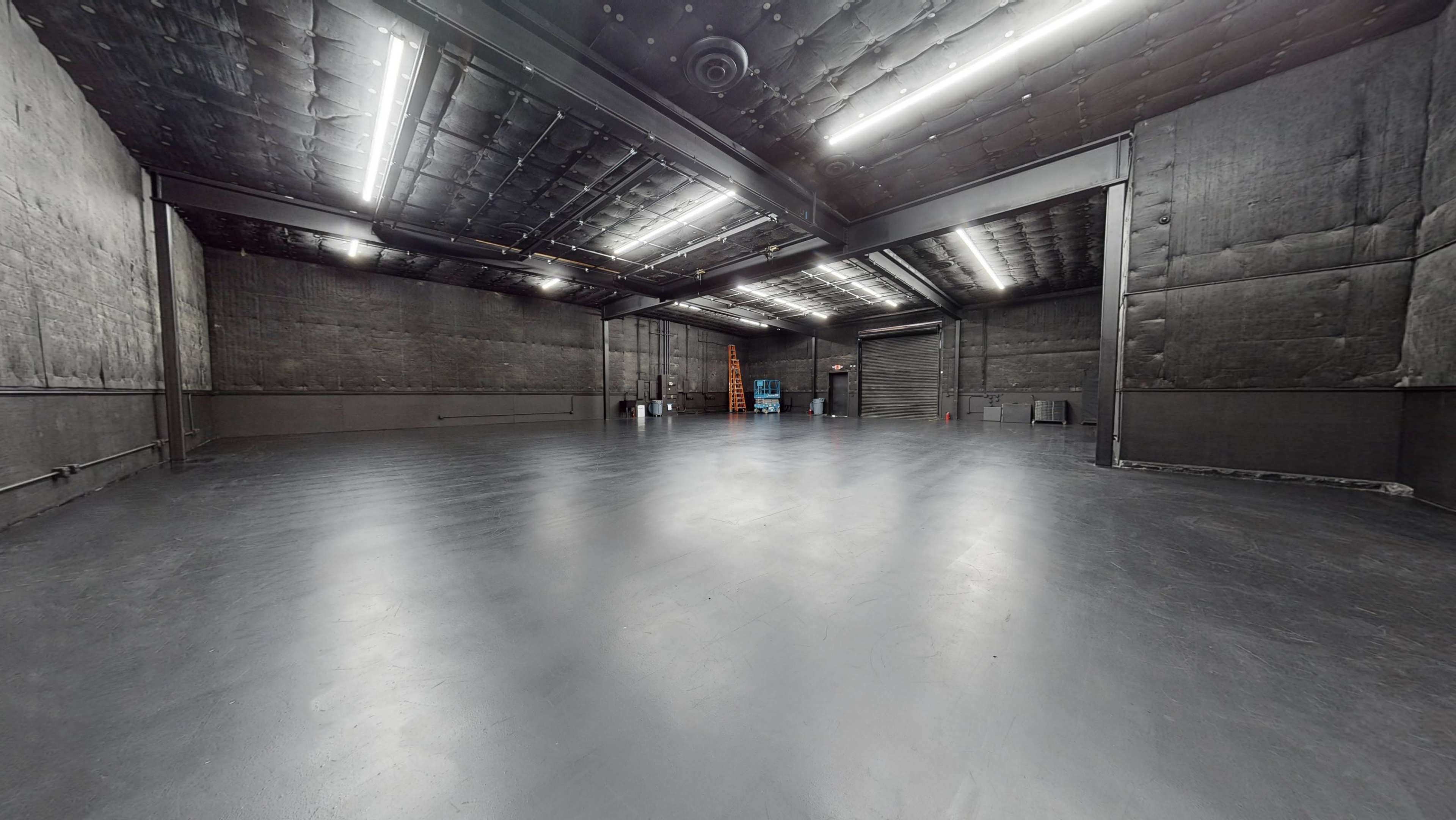 The image shows a large, empty warehouse space with dark walls and a smooth gray floor, illuminated by overhead fluorescent lights.