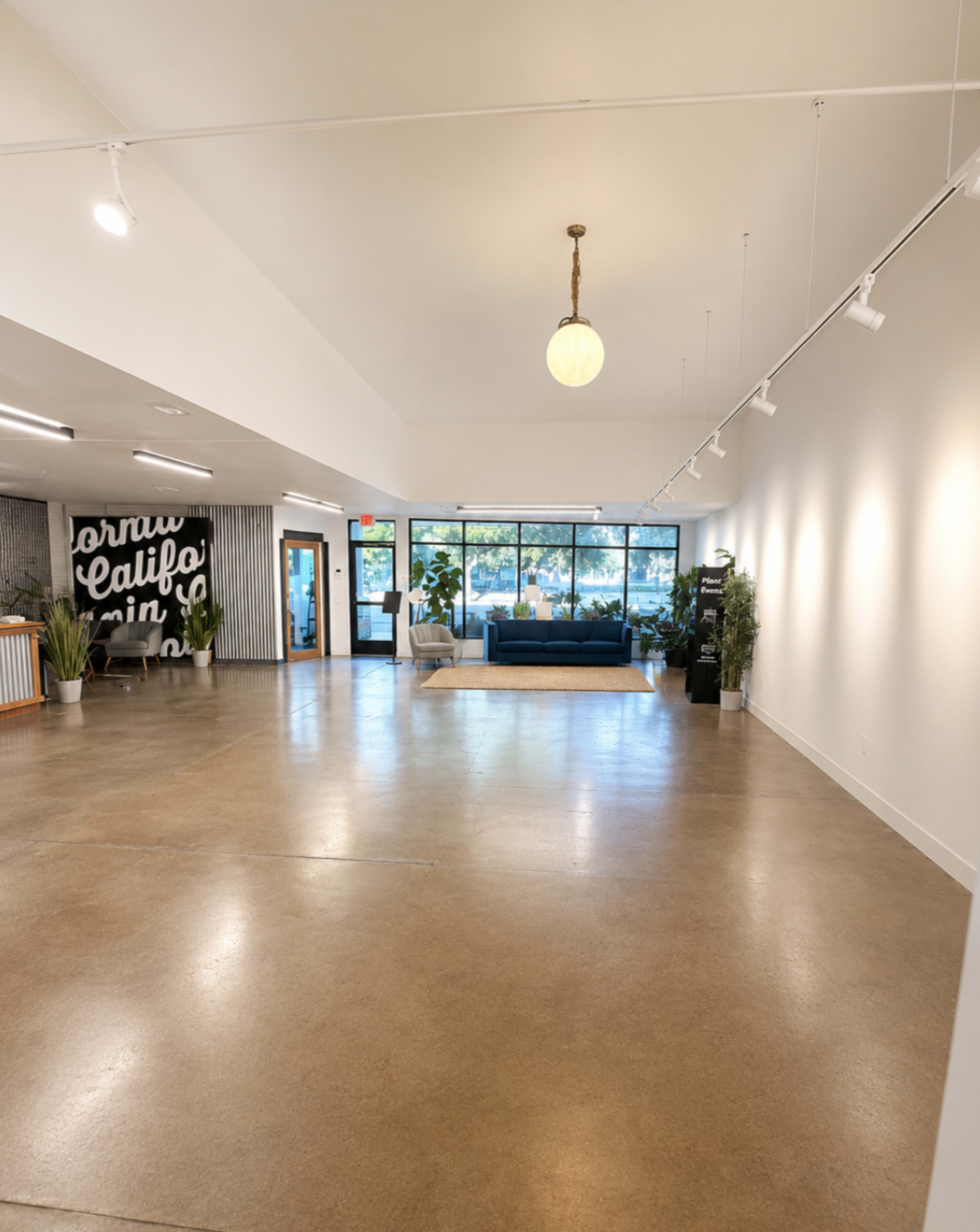 The image shows a spacious, well-lit interior with polished concrete flooring, a blue couch, large windows, and a black and white mural on one wall.