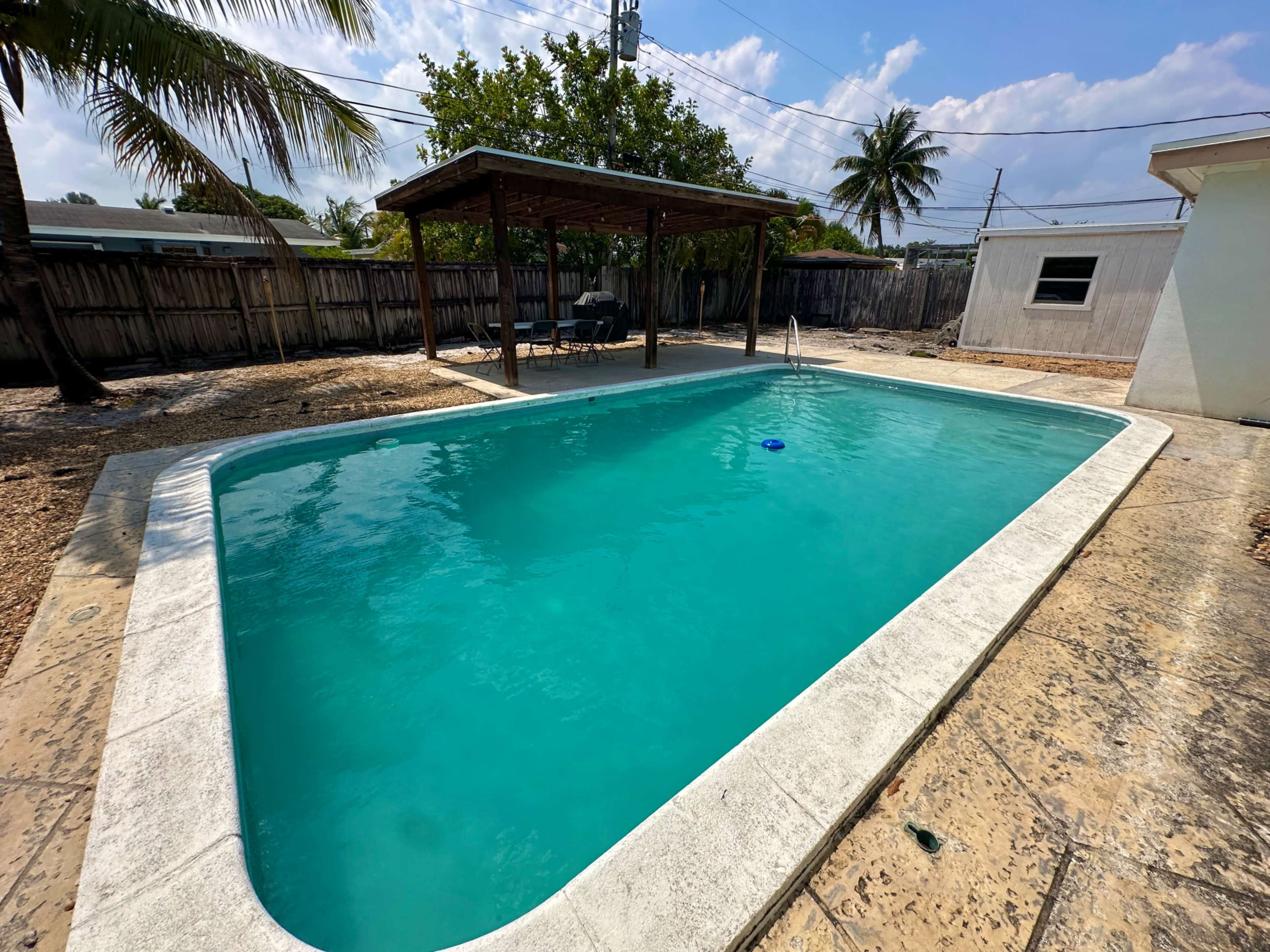 A clear swimming pool surrounded by a stone deck and a shaded pergola with chairs in a tropical setting.