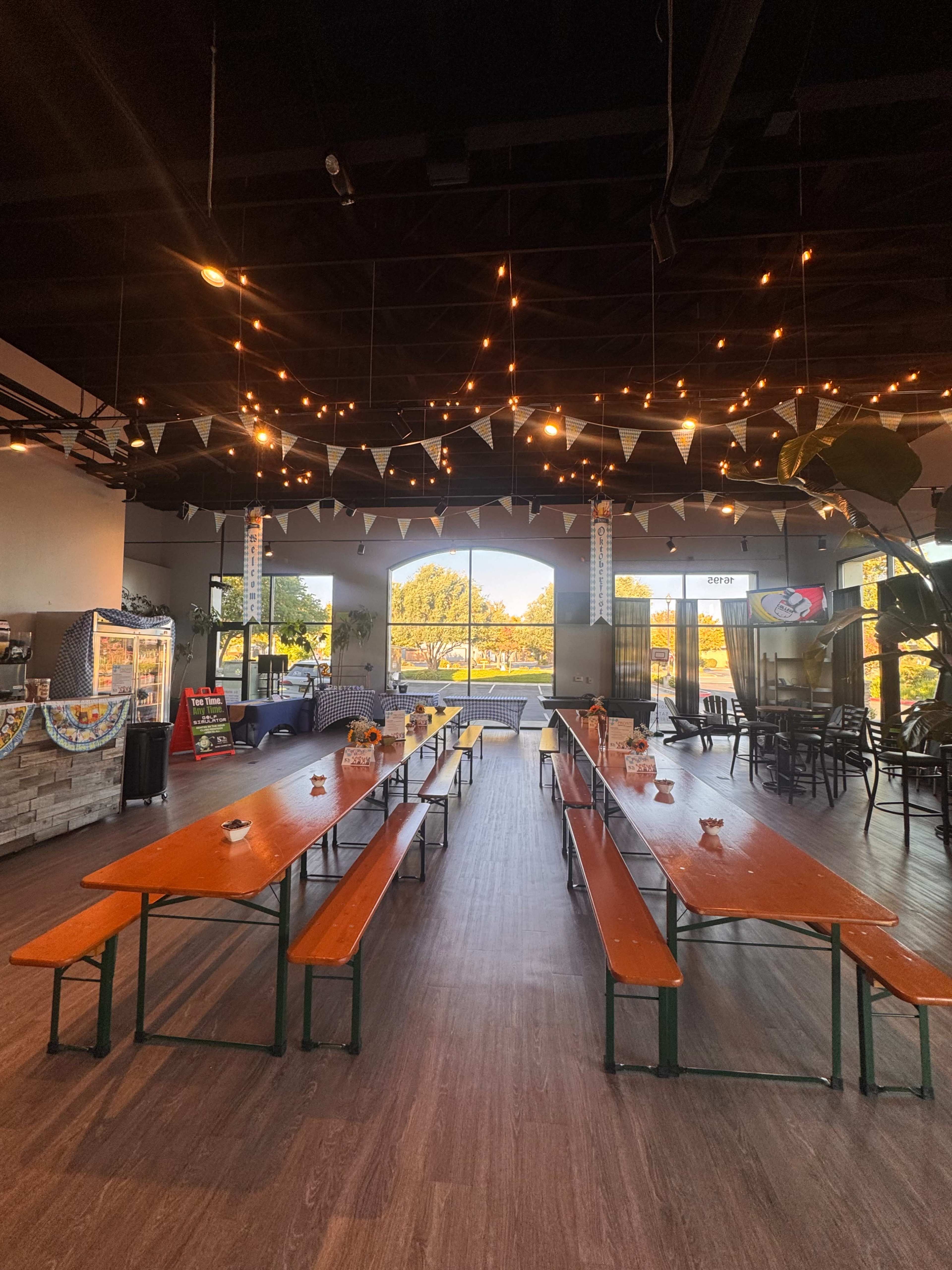 The image shows a spacious dining area in a café or restaurant featuring long wooden tables and benches, with string lights and decorative bunting overhead.