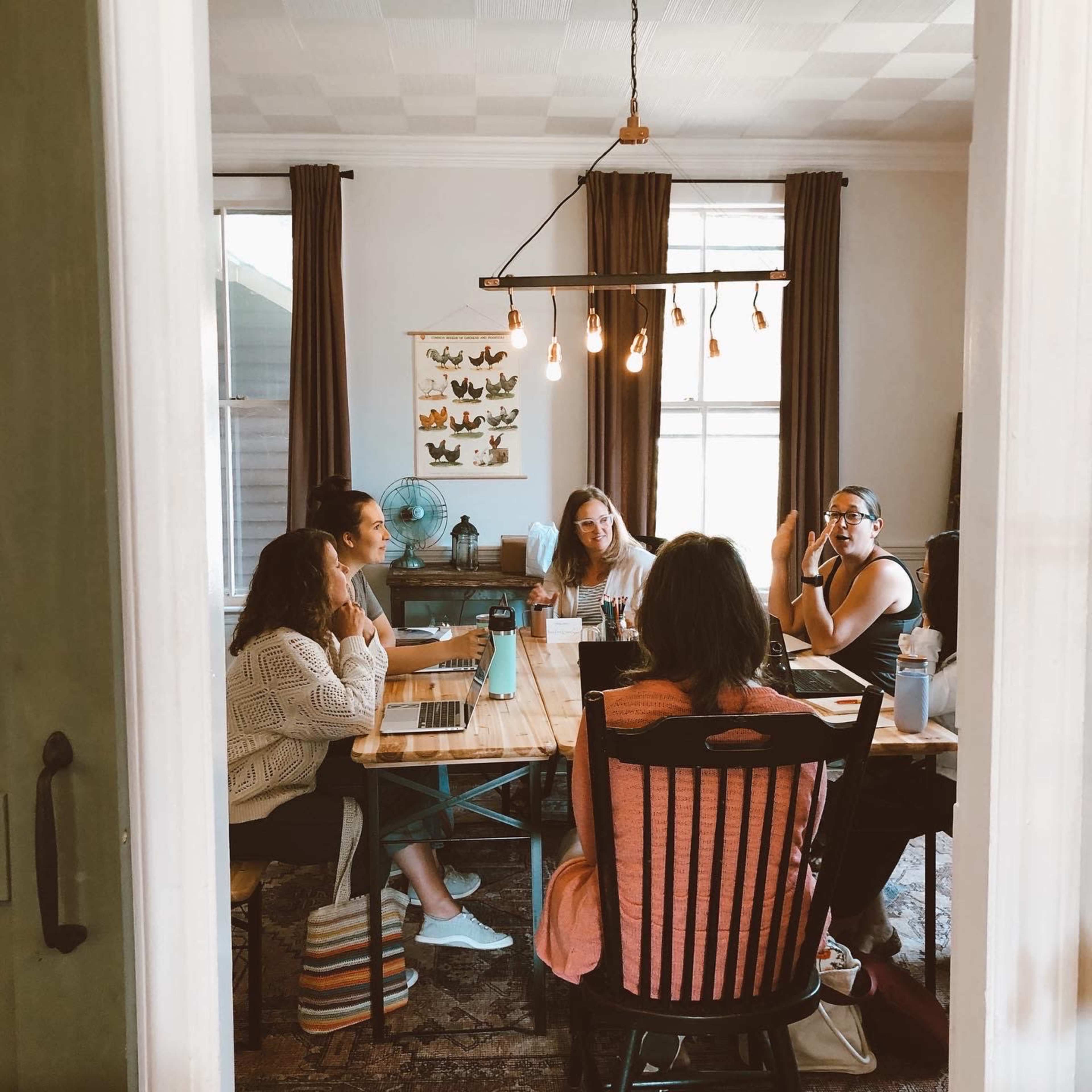 A group of six people sits around a wooden table in a well-lit room, engaged in a discussion with laptops and water bottles in front of them.