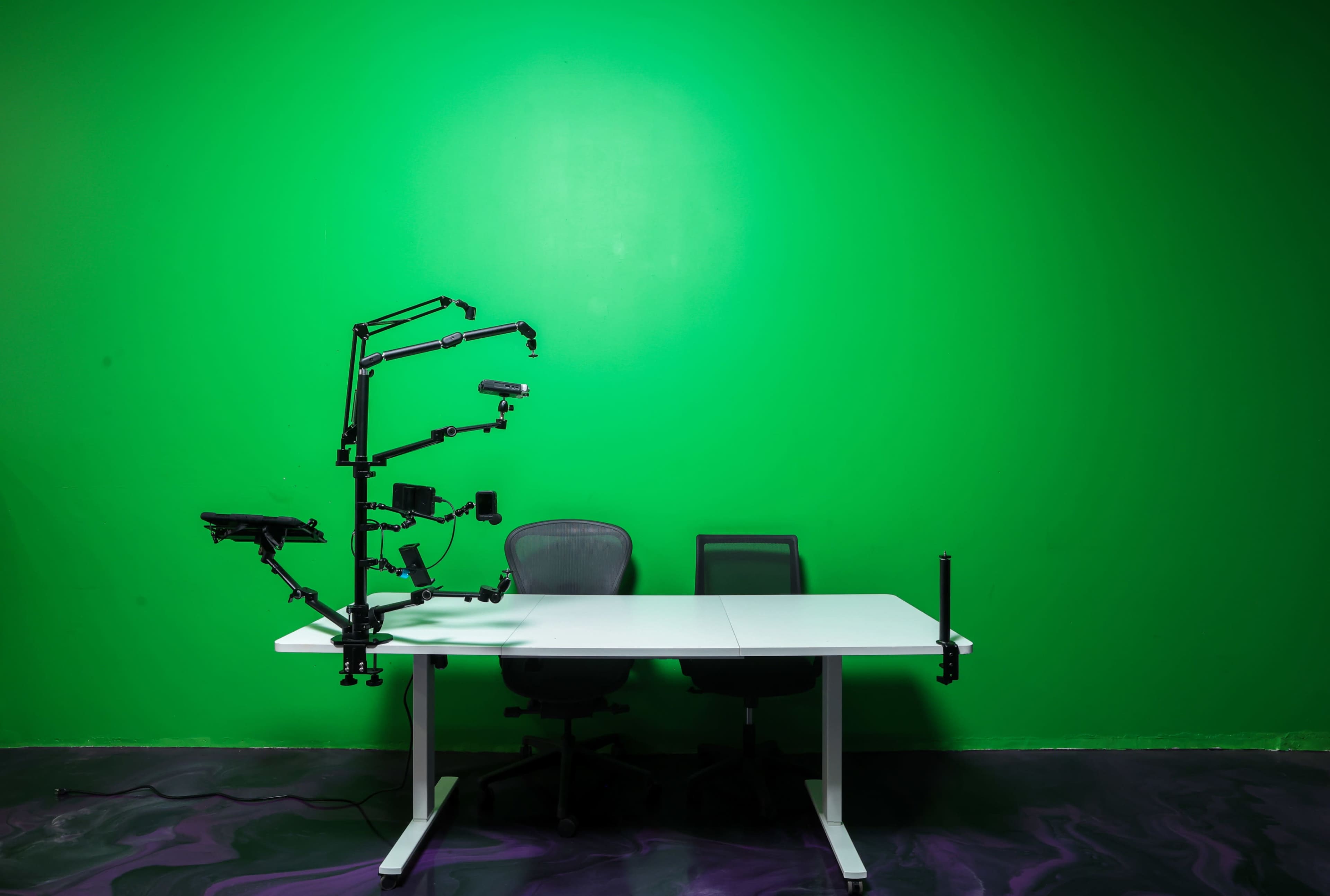 A green screen serves as the backdrop for a white desk with two chairs and an equipment arm mounted on one side.