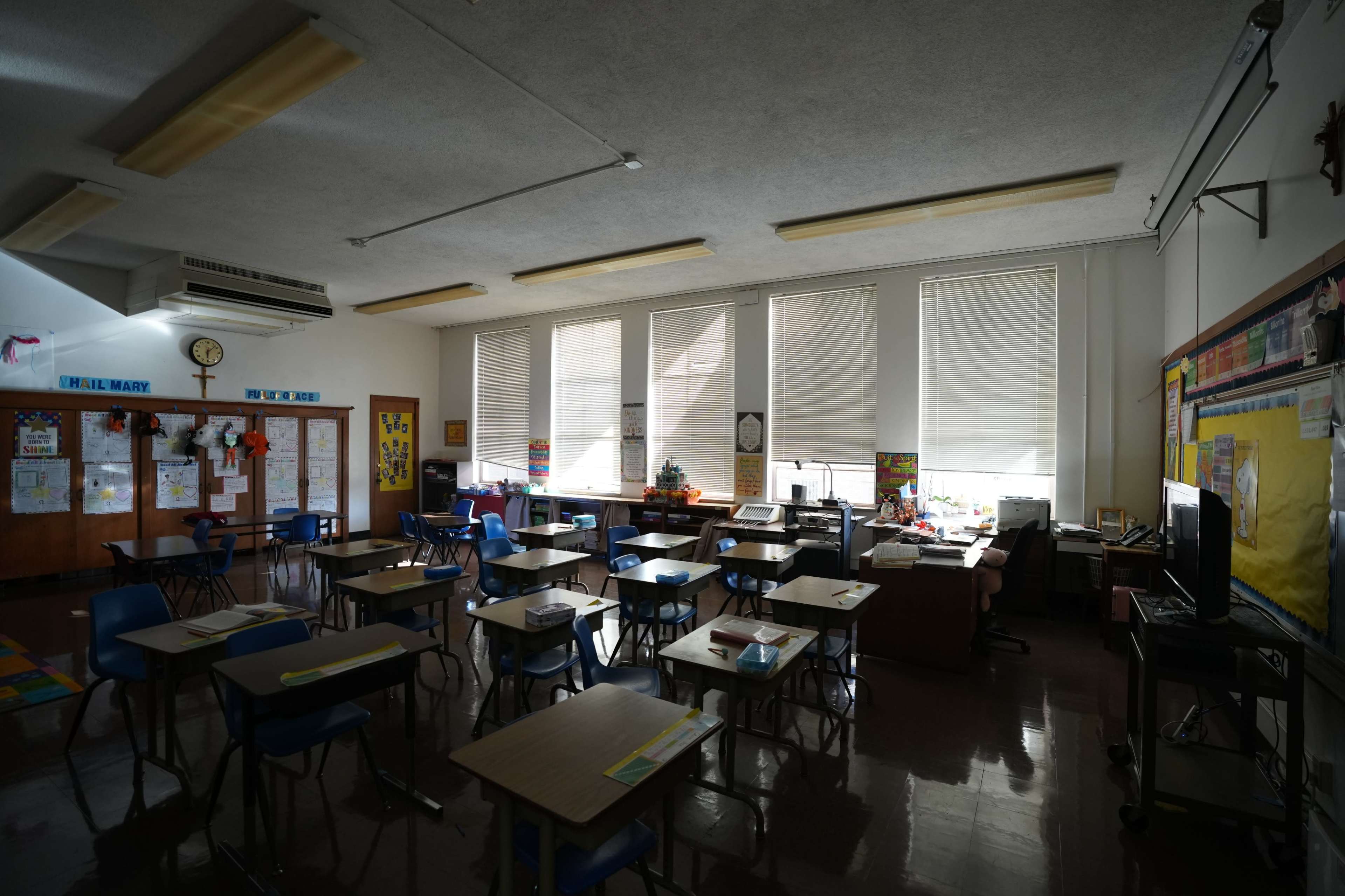 The image shows a nearly empty classroom with rows of desks, a teacher's desk, and large windows with blinds partially closed.
