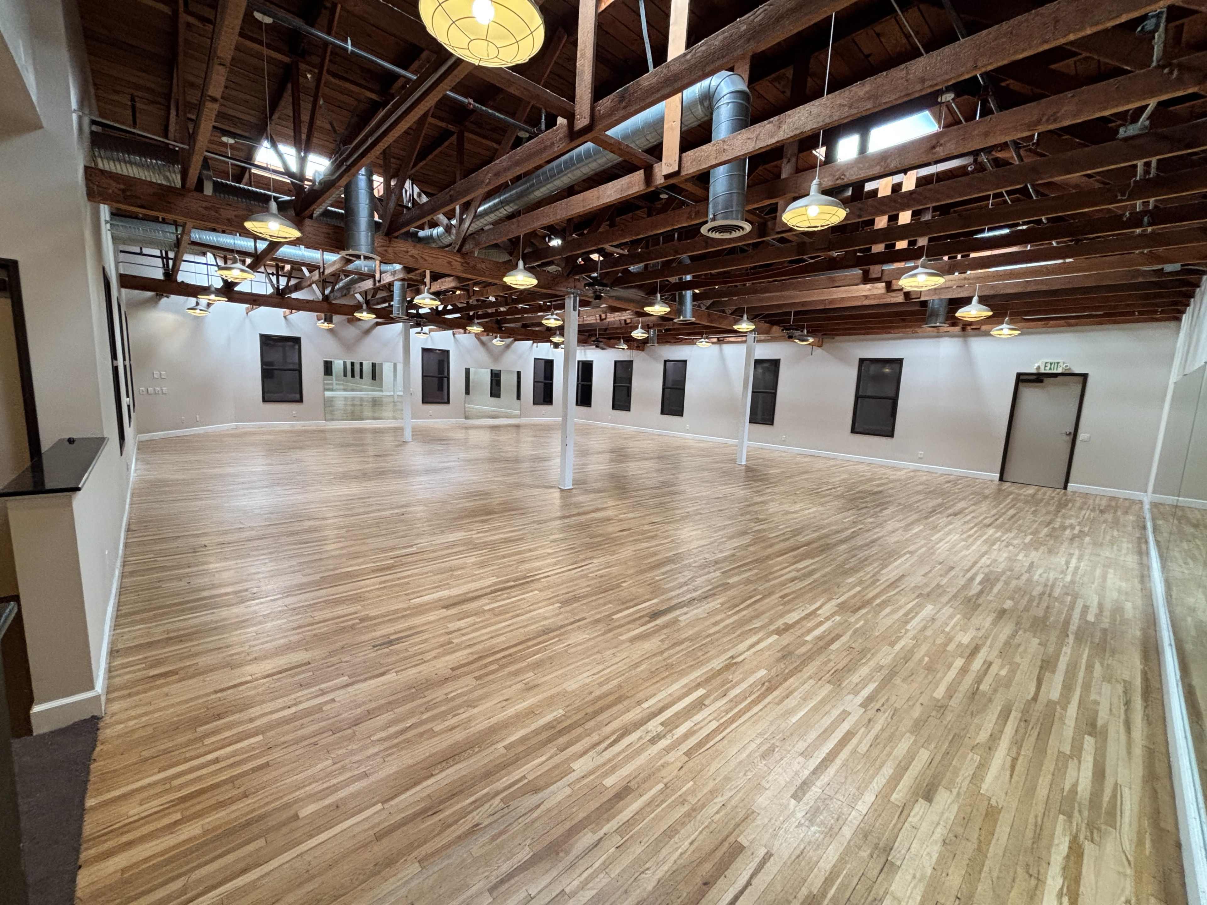 The image depicts a spacious studio with hardwood floors, high wooden ceilings, and multiple windows providing natural light.
