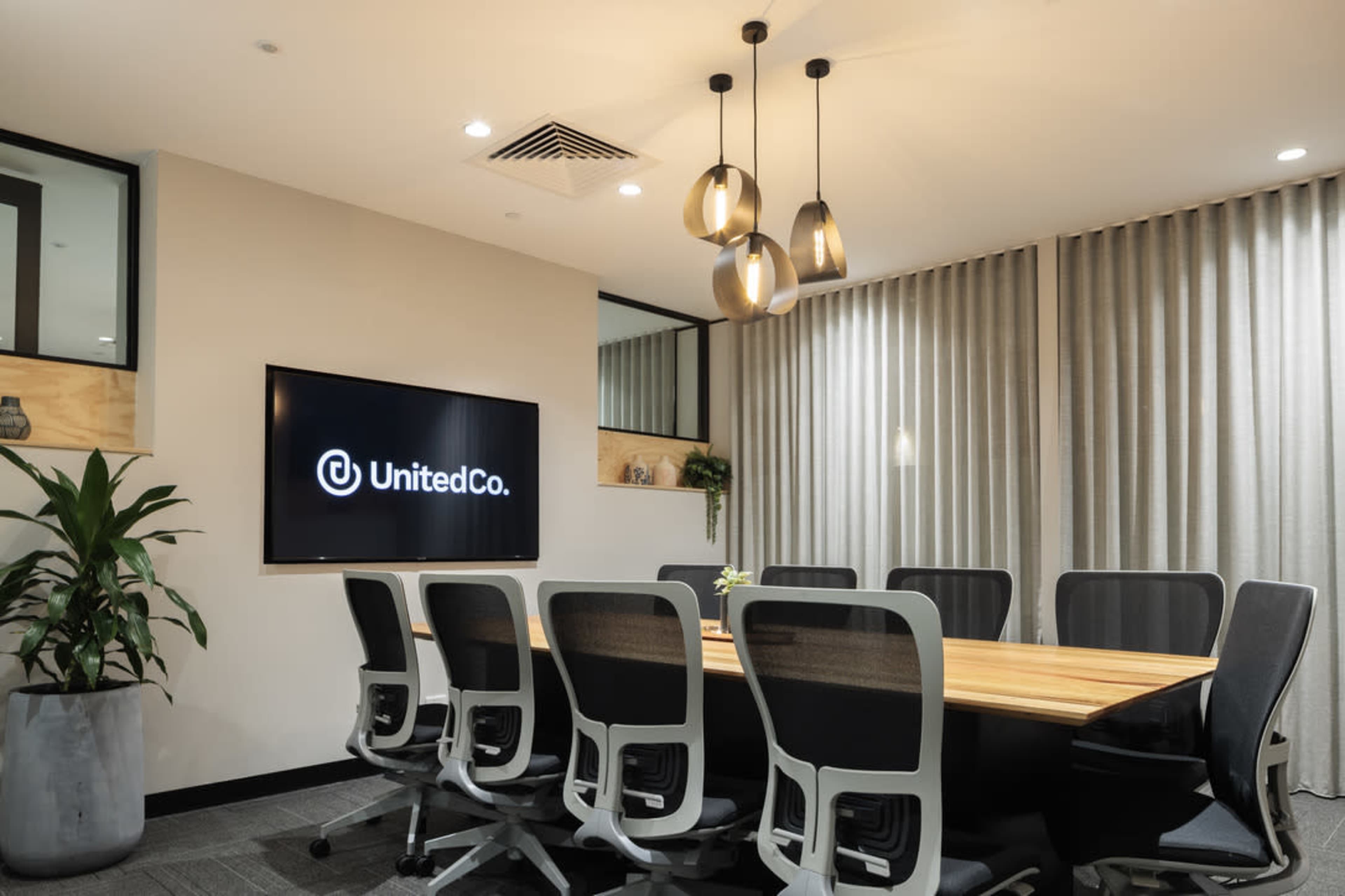 A modern conference room features a long wooden table surrounded by ergonomic chairs, with a large screen displaying the UnitedCo logo on the wall.