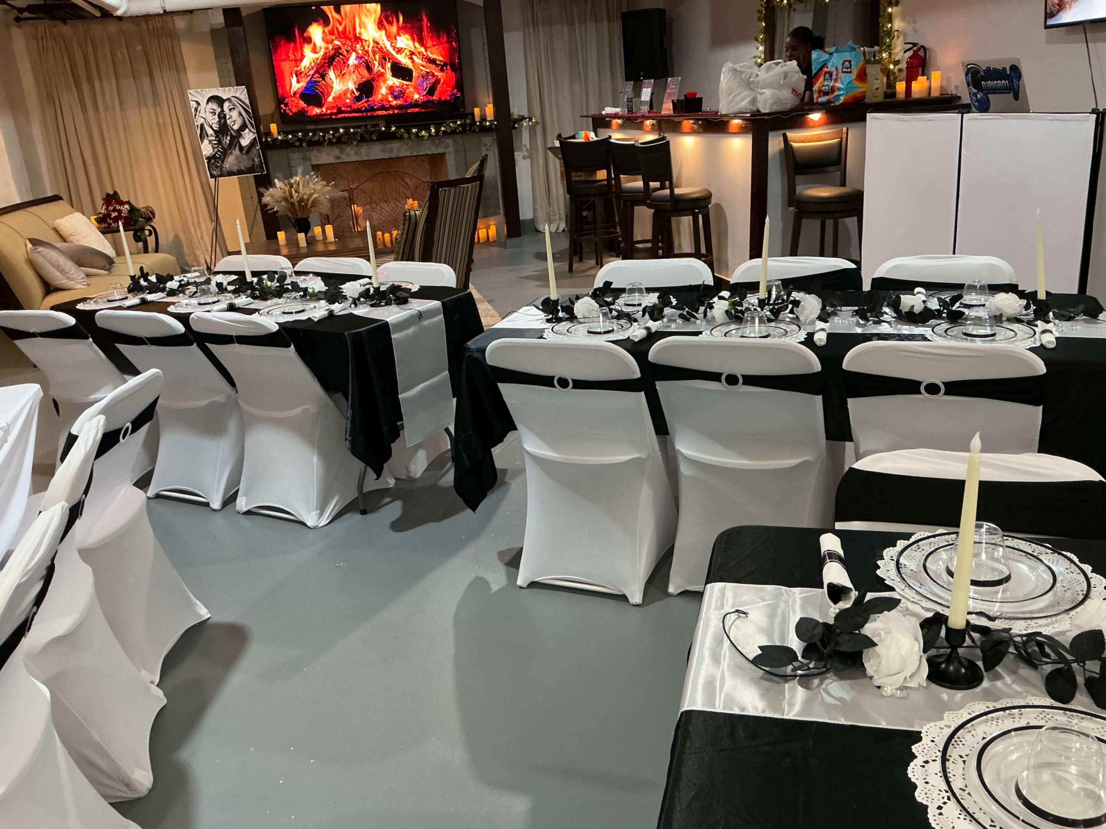 The image shows a decorated event space with tables set in alternating black and white linens, complete with elegant place settings and candles, and a bar area in the background.