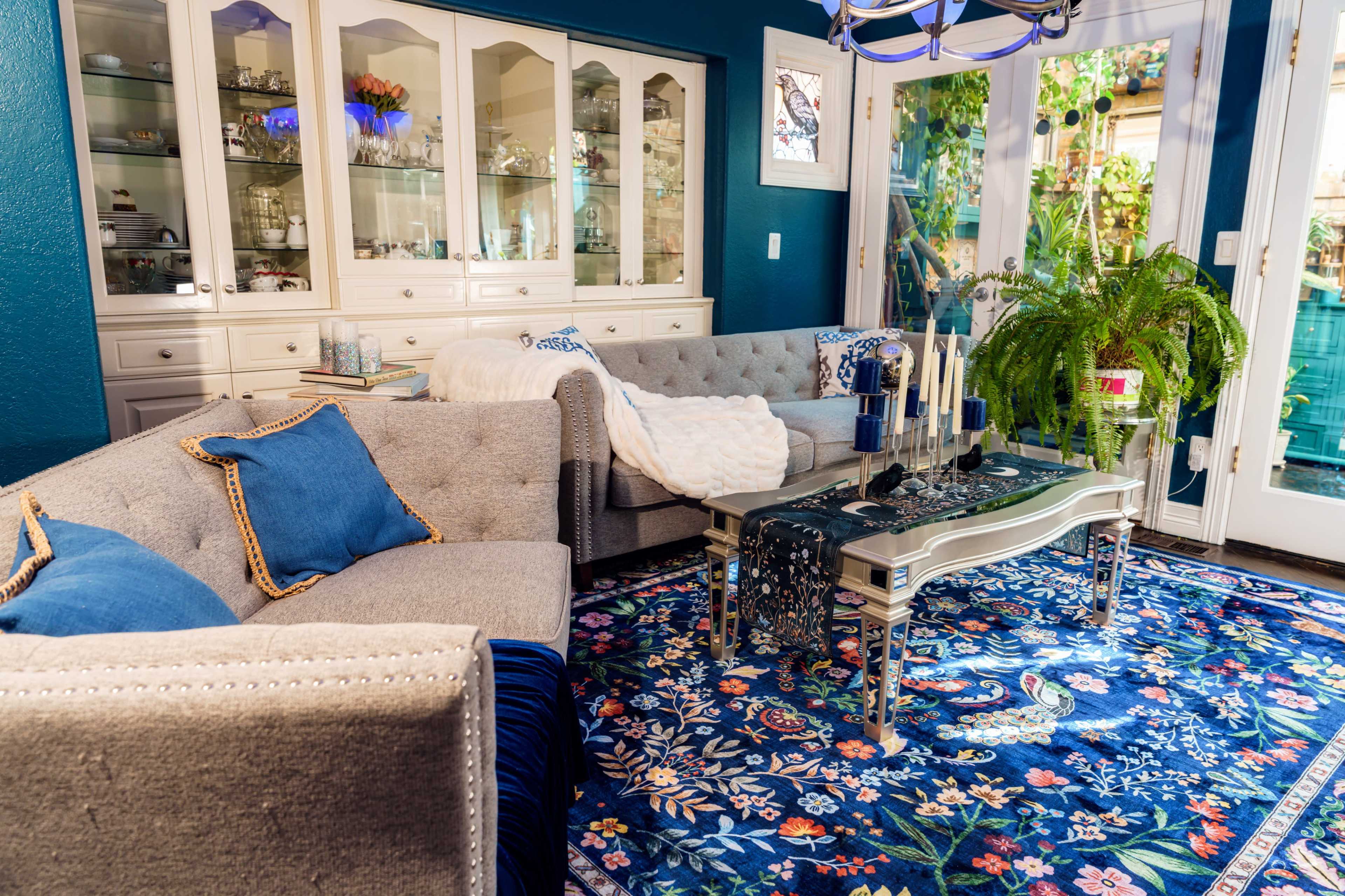 The image shows a living room with a gray sectional sofa, a glass coffee table, and a colorful area rug, surrounded by a white cabinet and large glass doors leading to a patio.