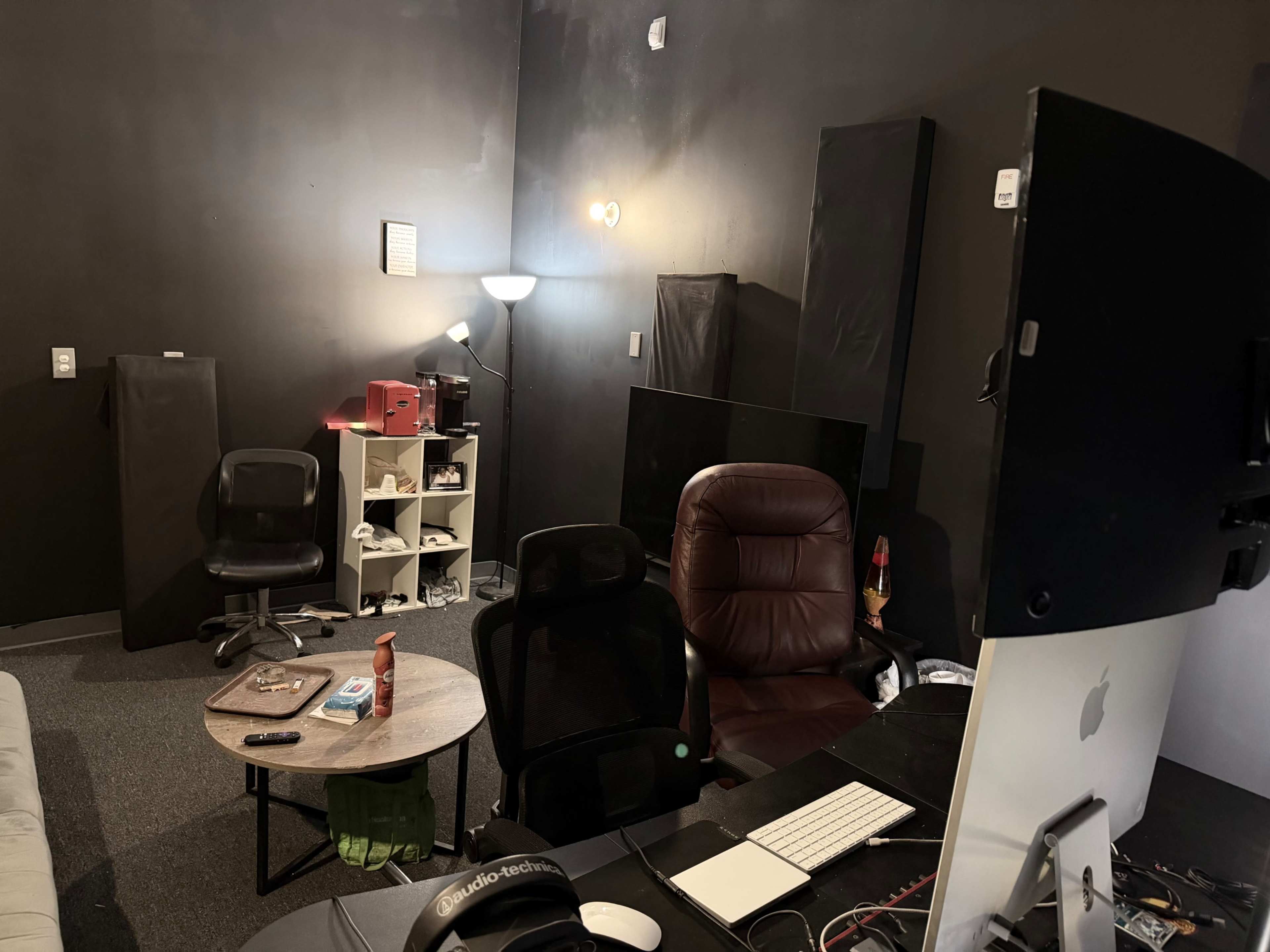 The image shows a dimly lit room with two office chairs, a small round table, a desk with a computer monitor, and several black acoustic panels on the walls.