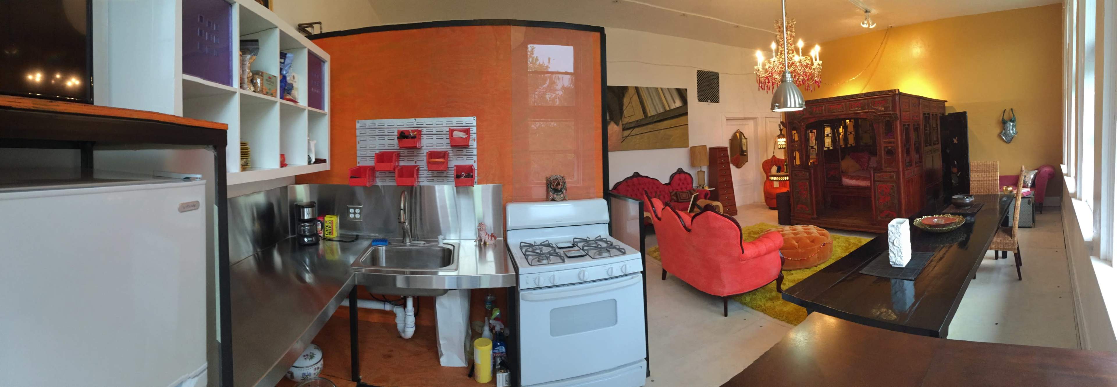 A panoramic view of a brightly colored kitchen and living area, featuring a stainless steel sink, a white stove, and a cozy seating arrangement with an ornate cabinet in the background.