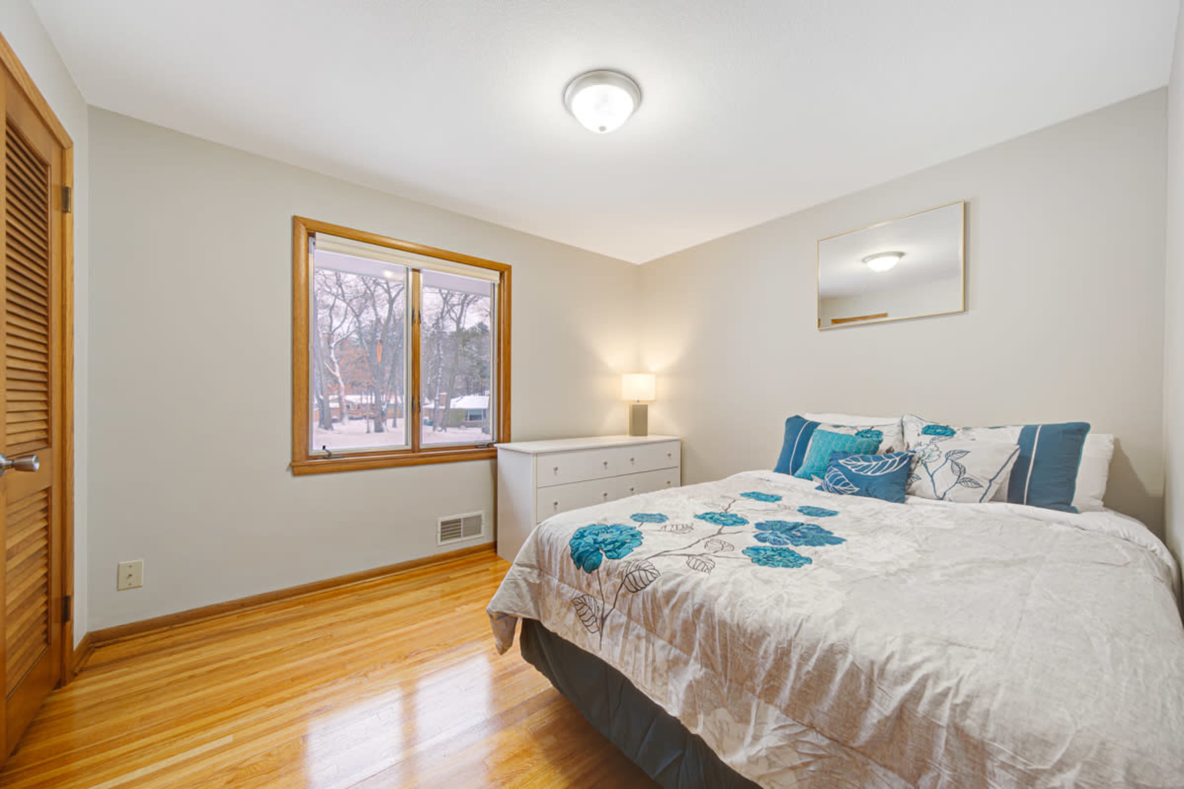 A well-lit bedroom features a bed with a floral-patterned comforter, a dresser, and a window overlooking a snowy landscape.