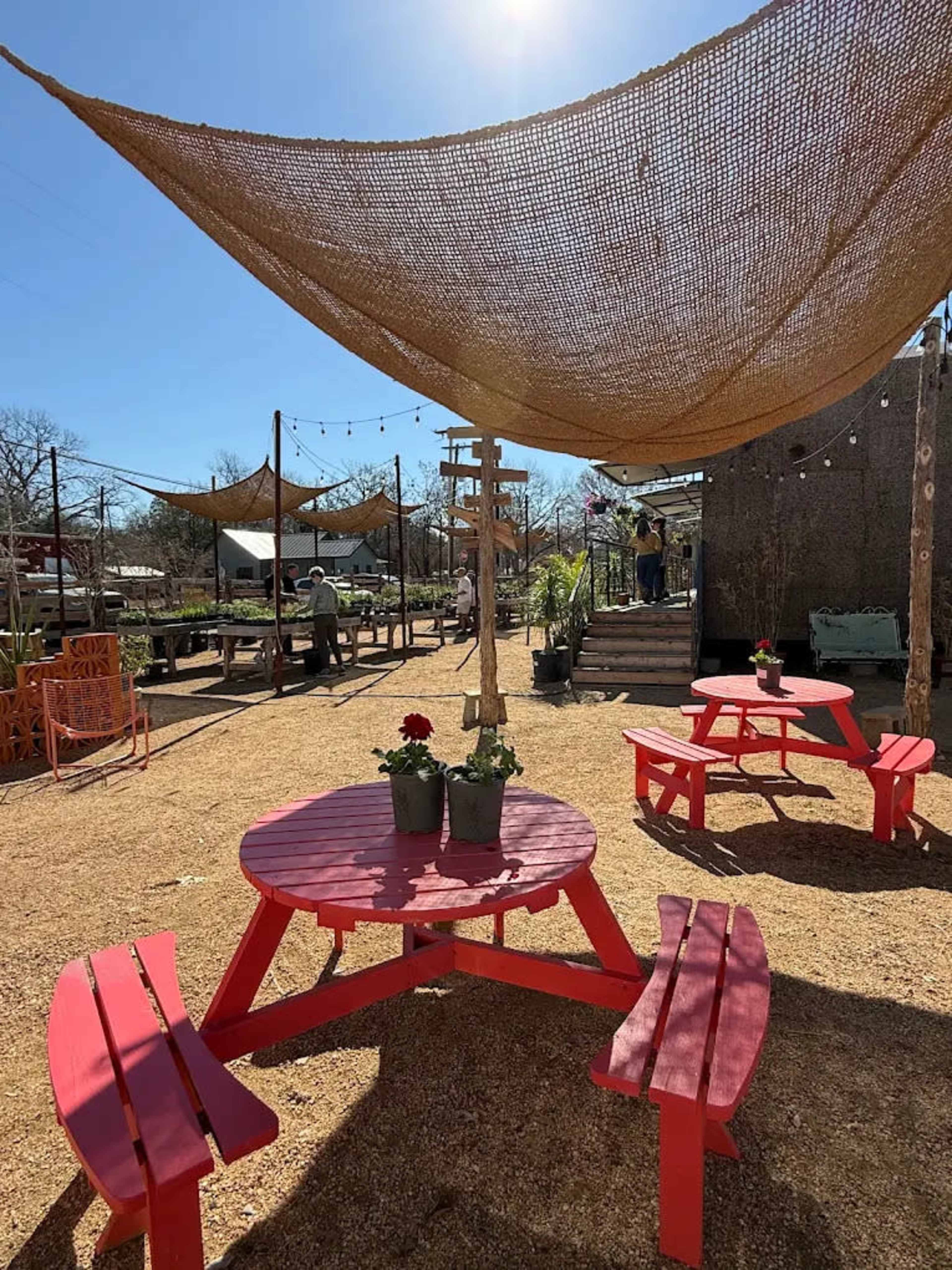 The image shows a sunny outdoor seating area with red picnic tables, planters with flowers, and a canopy overhead, surrounded by gravel and wooden structures.