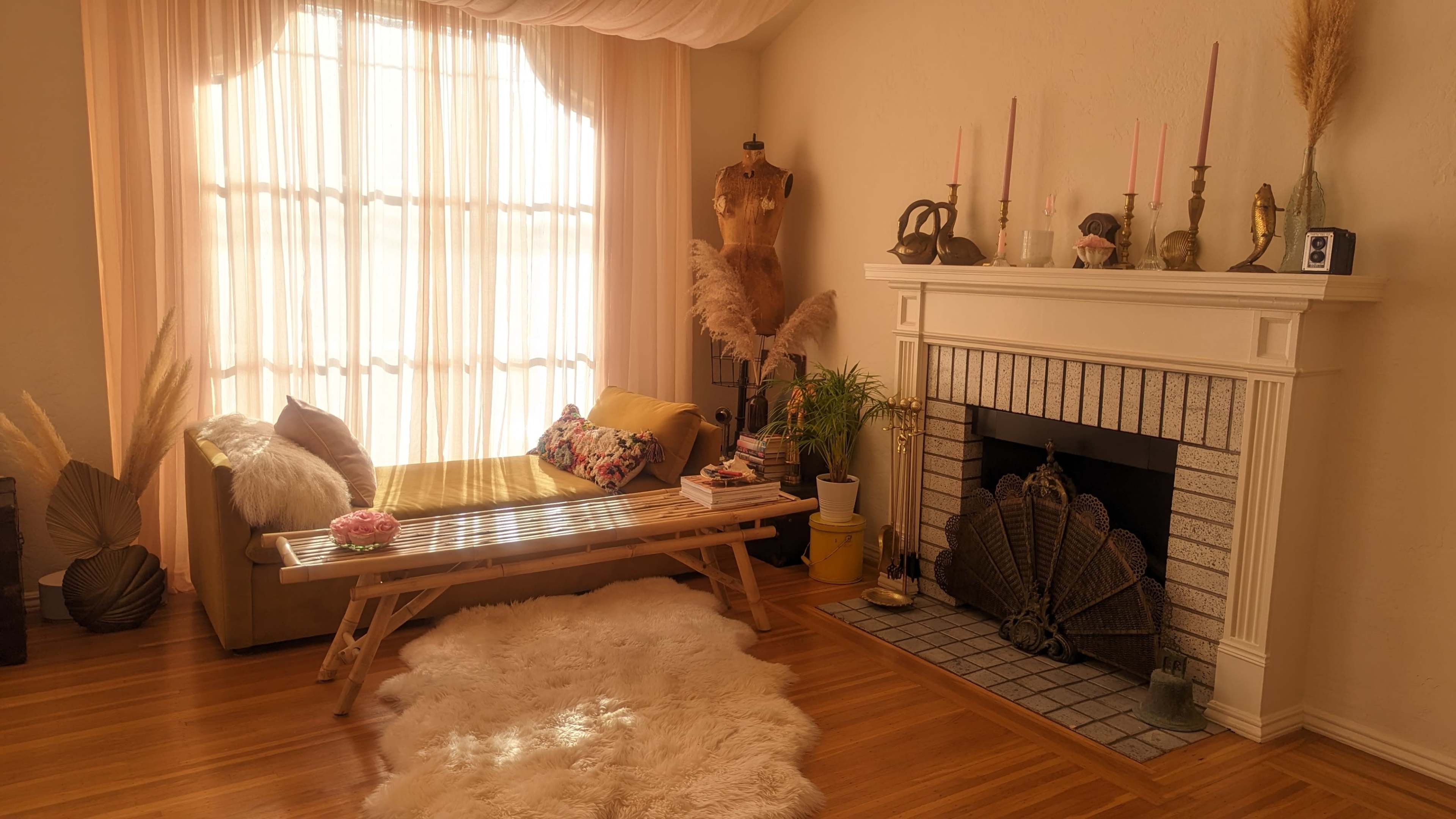 A cozy living room features a sofa, a coffee table, a decorative fireplace, and large windows with sheer curtains allowing natural light to fill the space.
