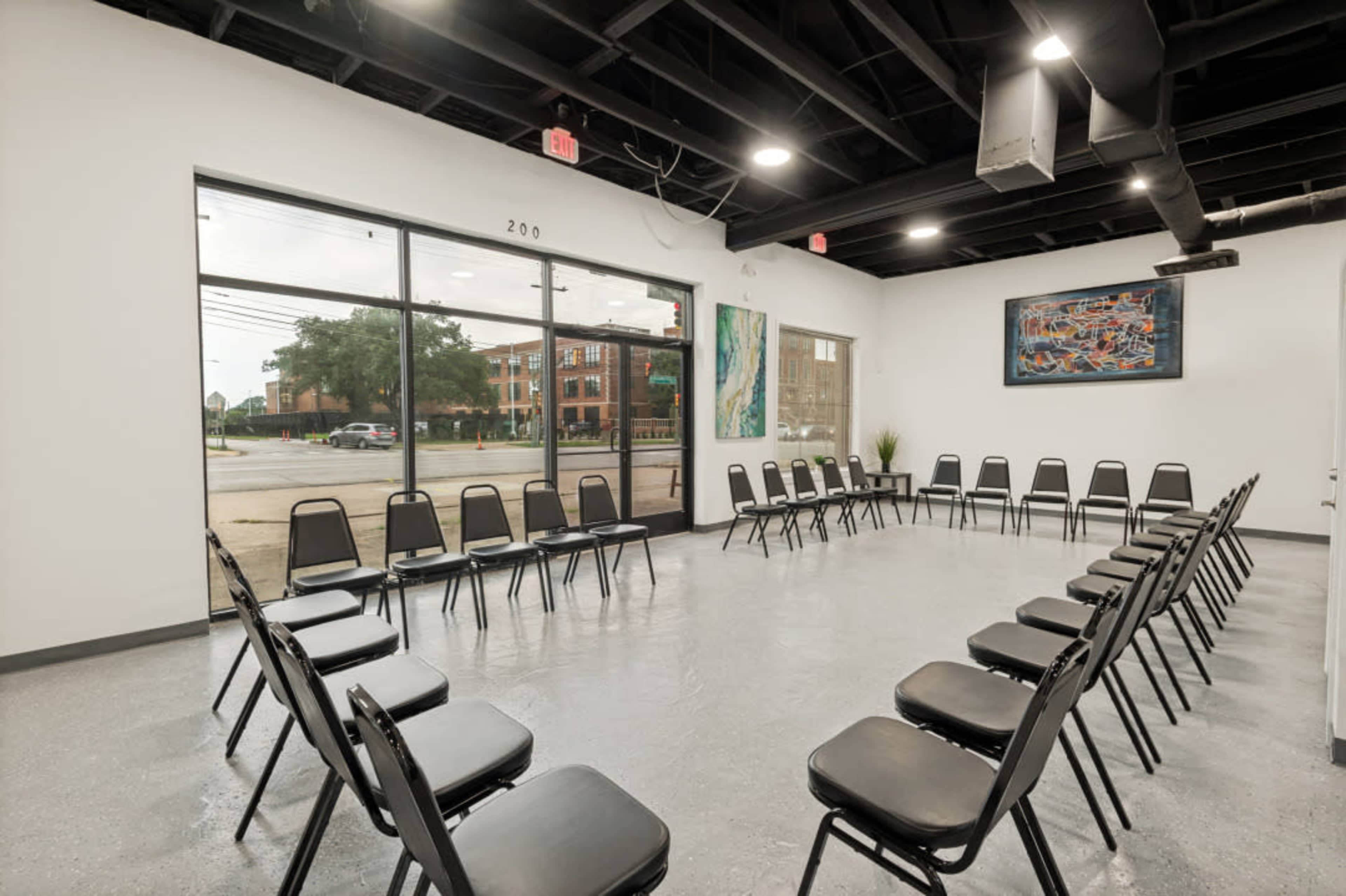 The image shows a spacious room with a circular arrangement of black chairs facing an open window, revealing an exterior view of a street and buildings.