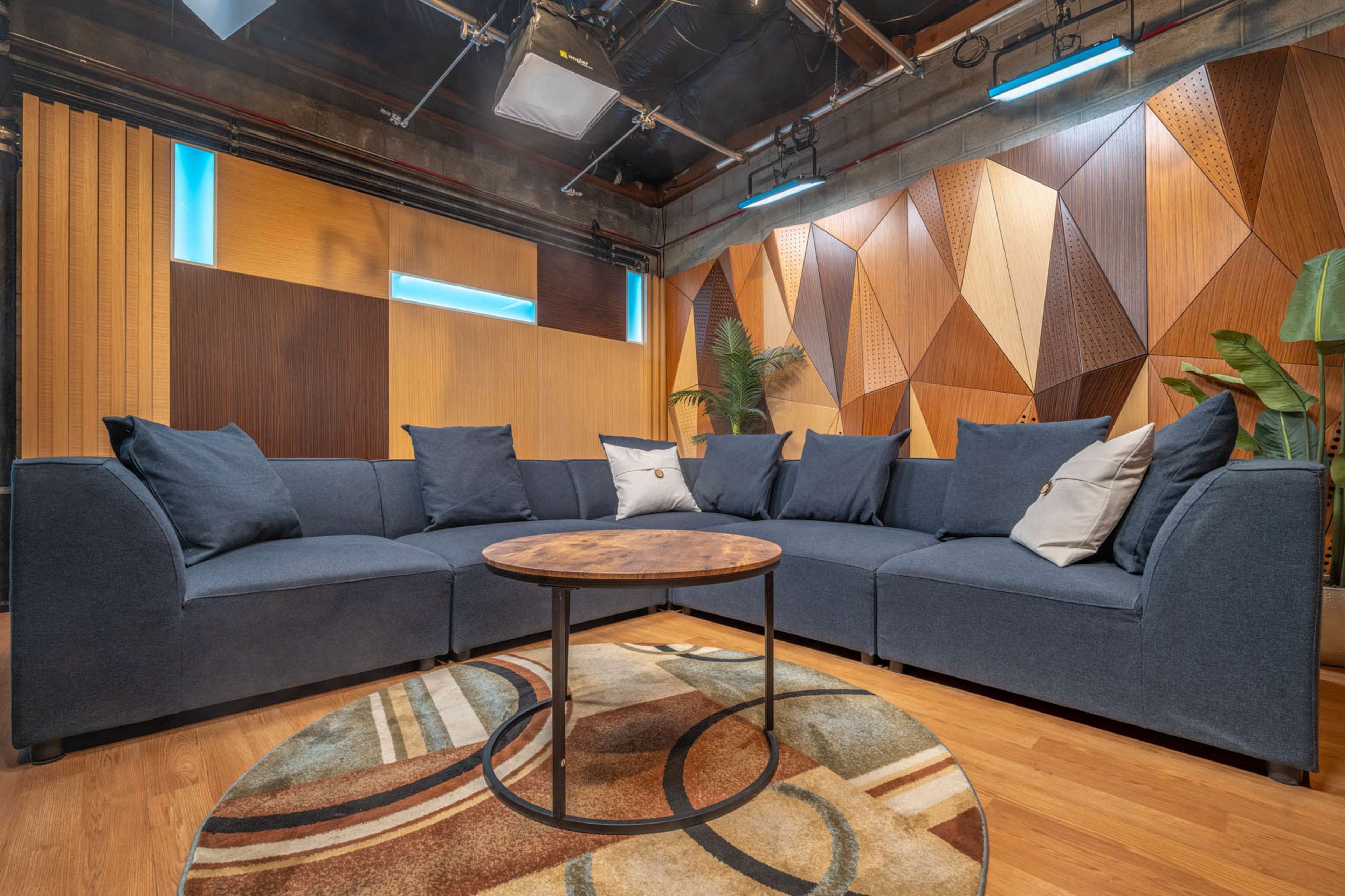 A modern sectional sofa with gray fabric and decorative pillows surrounds a circular wooden coffee table on a patterned rug in a well-lit studio with textured wooden panels on the walls.