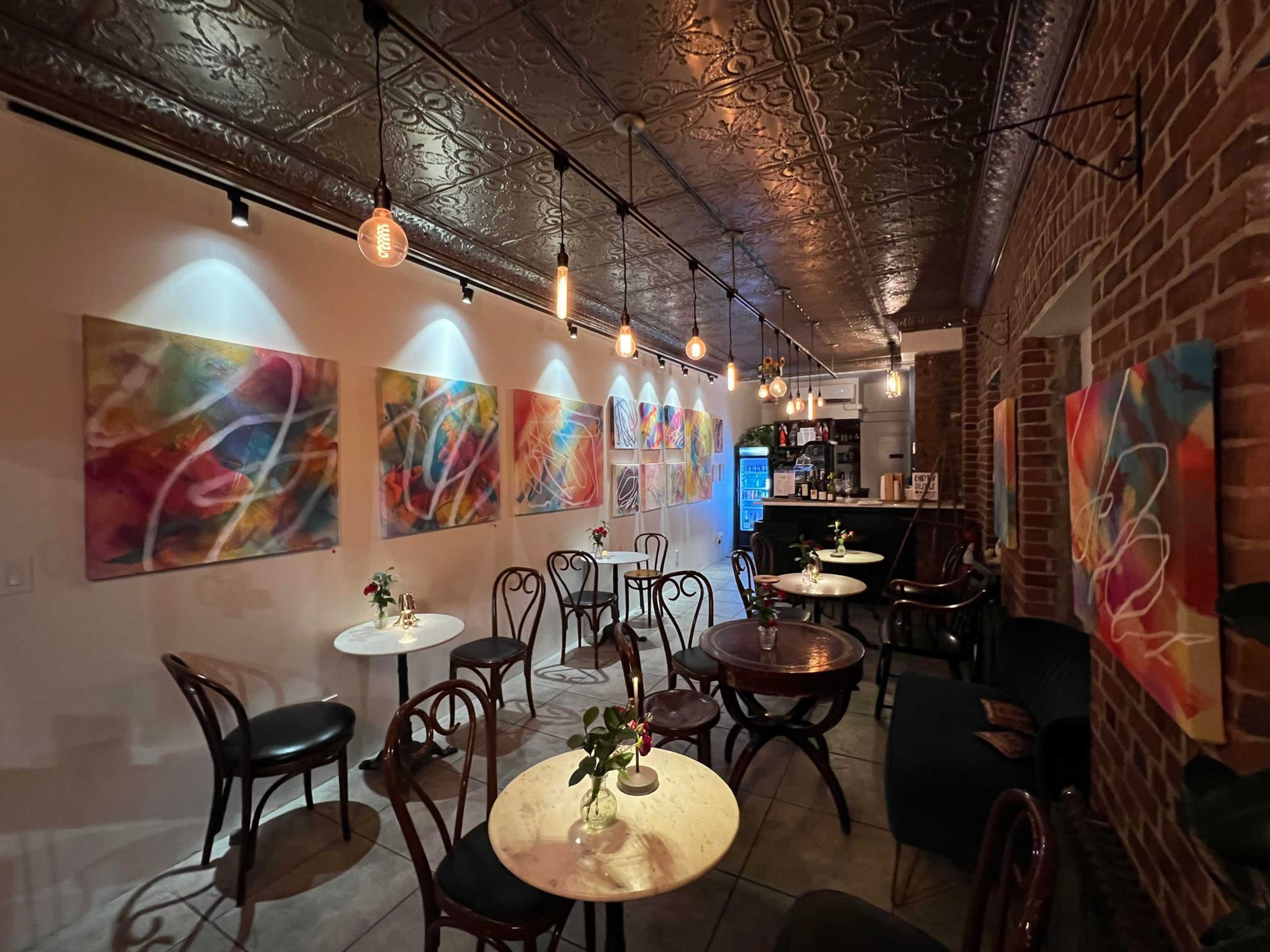 A cozy café interior with several small tables, wooden chairs, and vibrant abstract paintings on the walls, illuminated by hanging light bulbs.