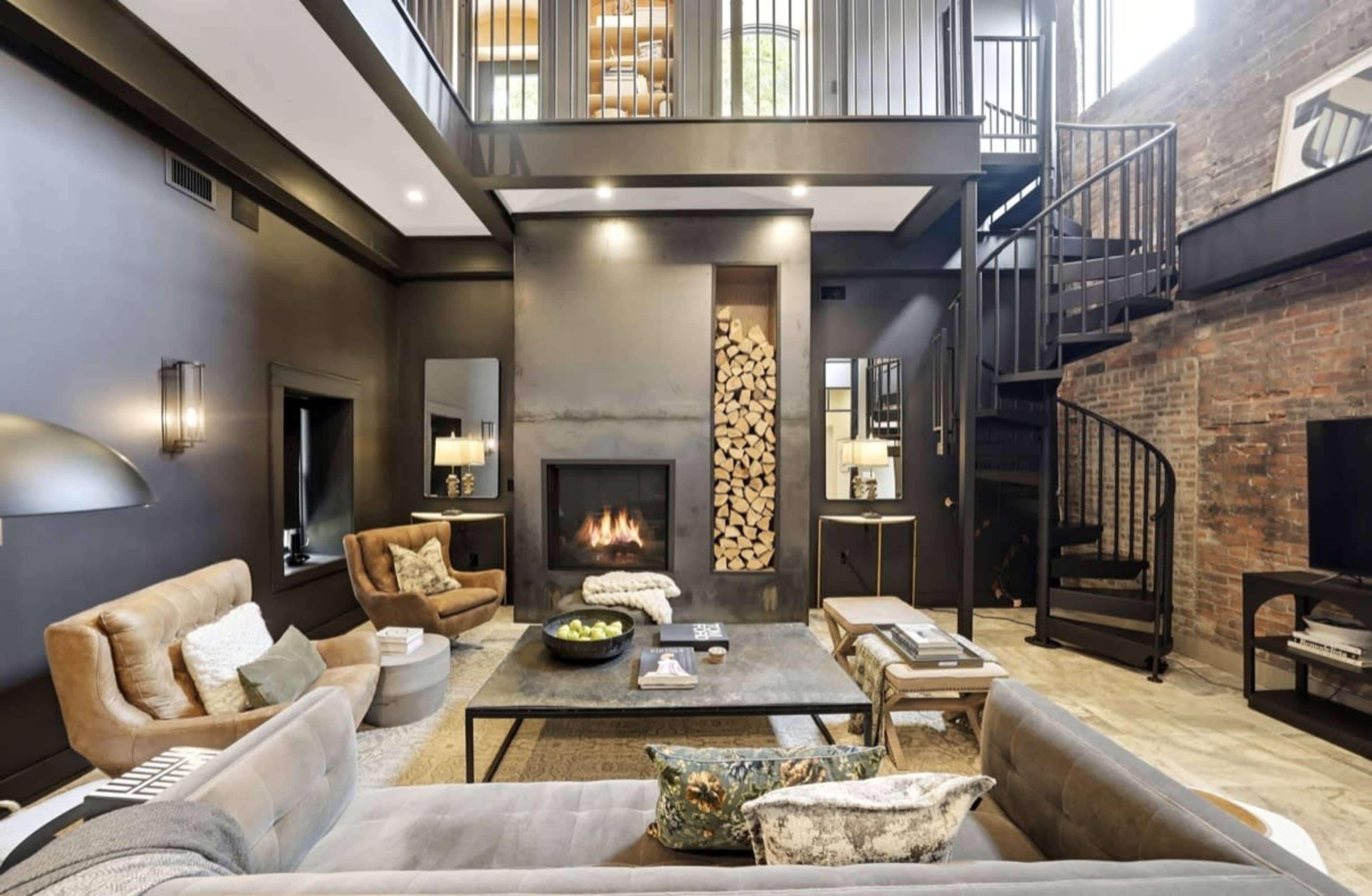 The living room features a modern design with a fireplace surrounded by stacked wood, plush seating, and a spiral staircase leading to a loft.