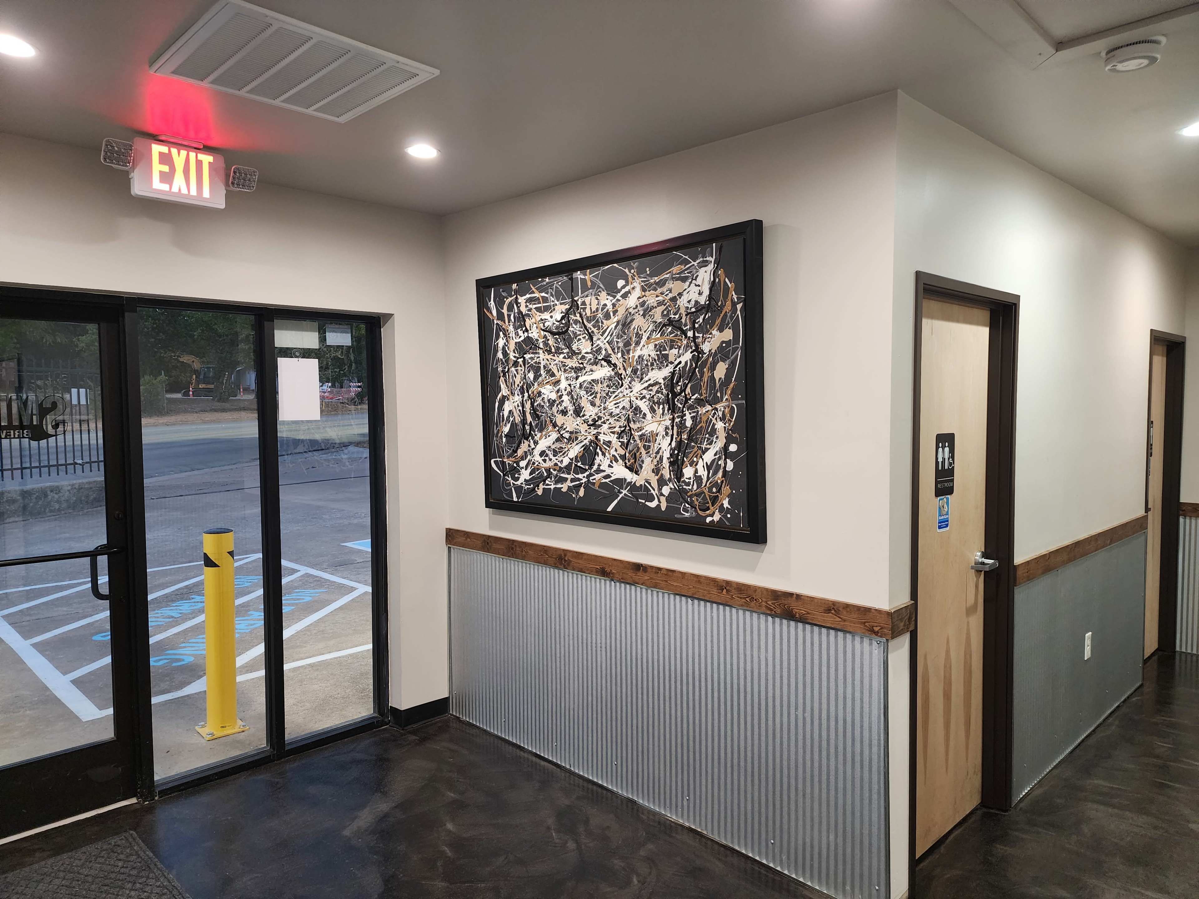 The interior of a building features a hallway with an exit door, a framed abstract painting on the wall, and an entrance to an office or room.