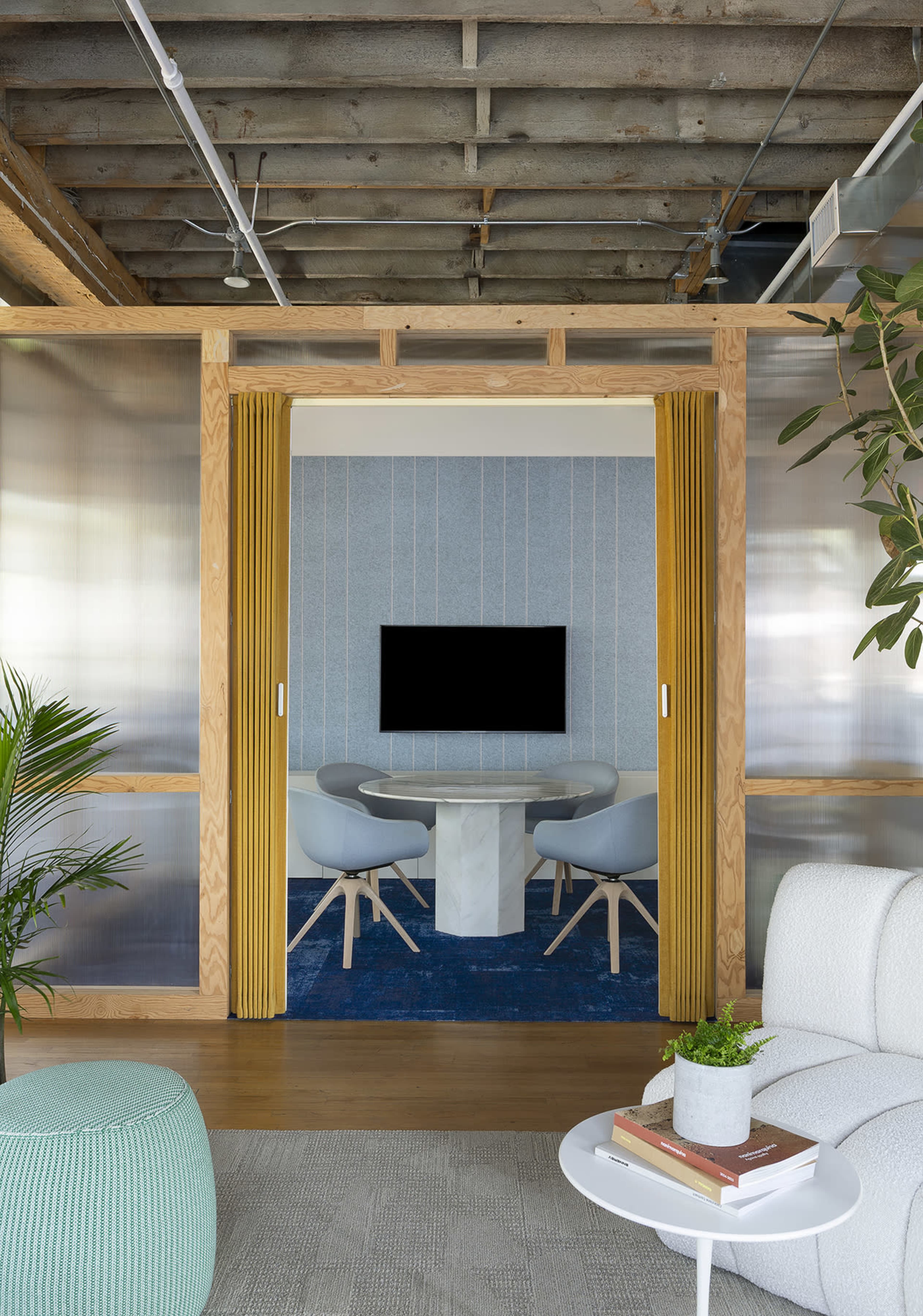 The image shows a modern office space featuring a small meeting area with two gray chairs, a round table, and a mounted television, framed by wooden elements and yellow curtains.