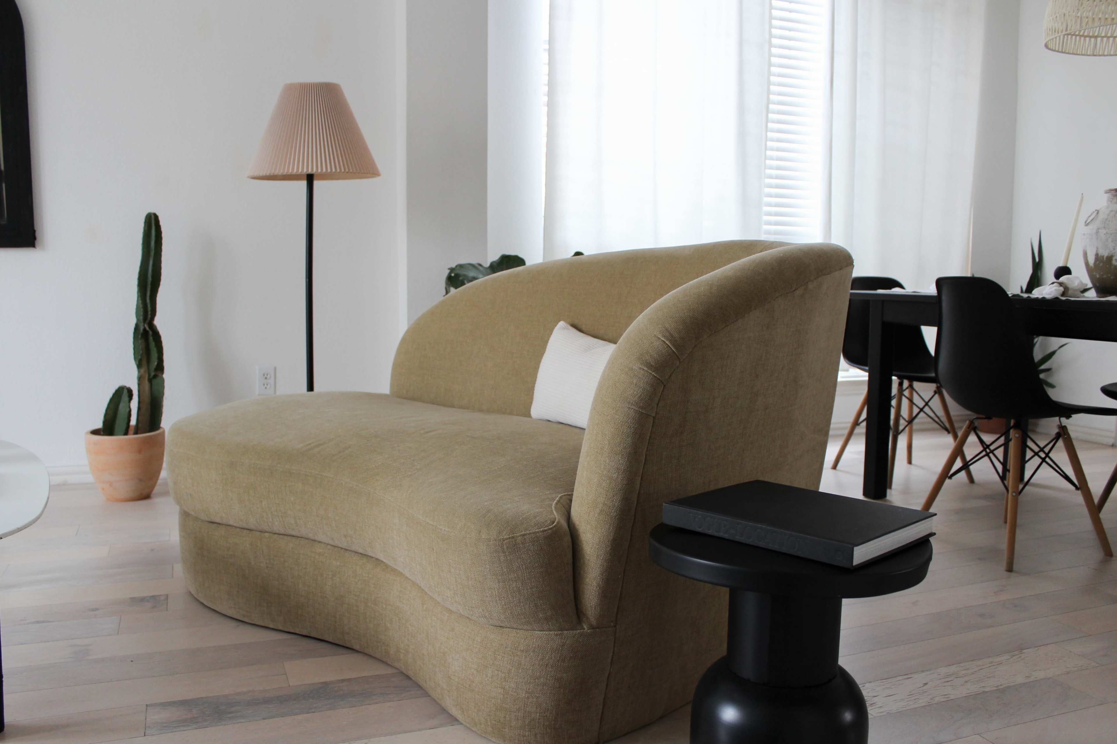 The image shows a beige curved sofa next to a floor lamp and a small black side table in a bright, minimalist interior space.