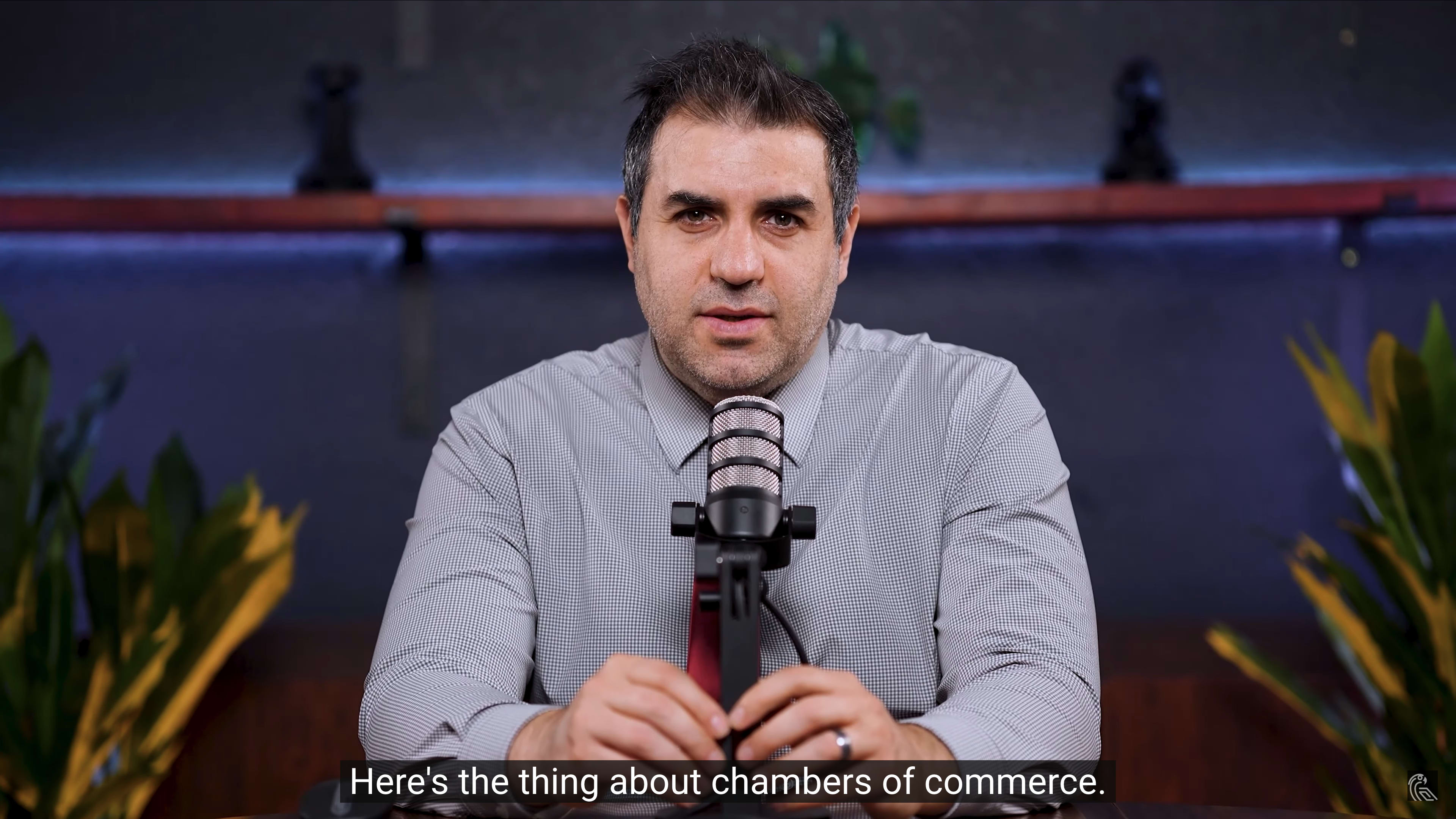 A man in a button-up shirt speaks into a microphone at a desk, with plants and decorative items in the background.
