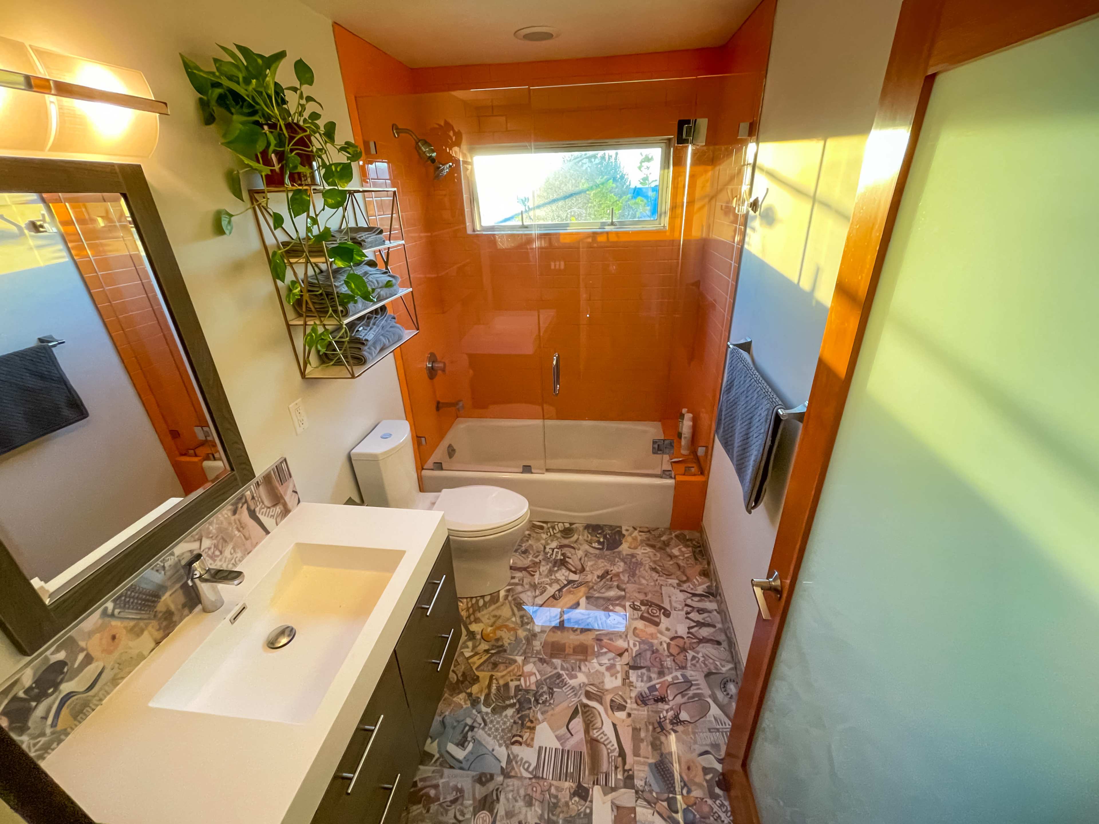 A modern bathroom featuring an orange accent wall, a shower-tub combo, a white sink with a dark cabinet, and a collage-style floor.