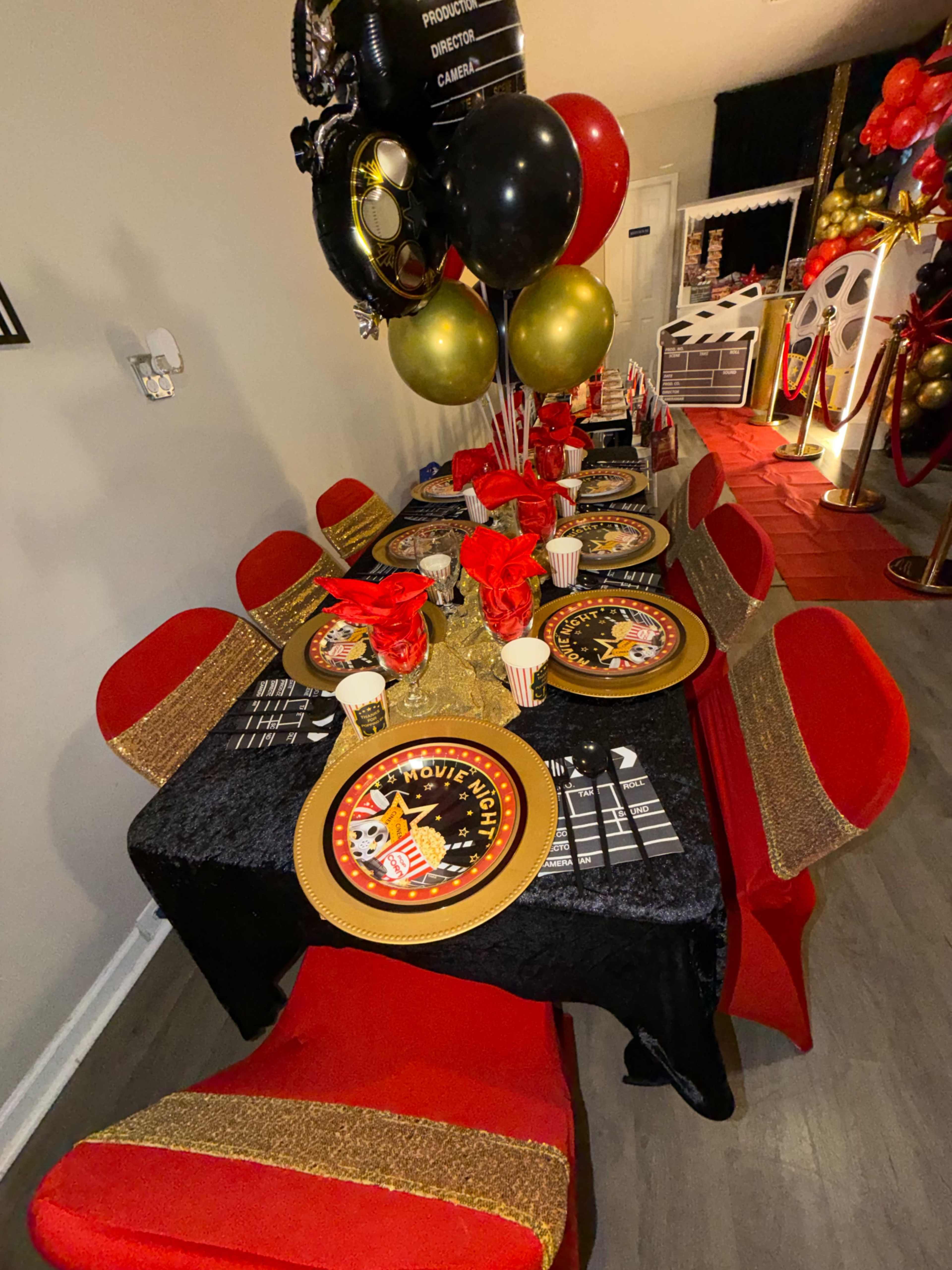 The image shows a neatly arranged party table decorated with black and gold tablecloths, red rose centerpieces, and themed plates, balloons, and party favors.