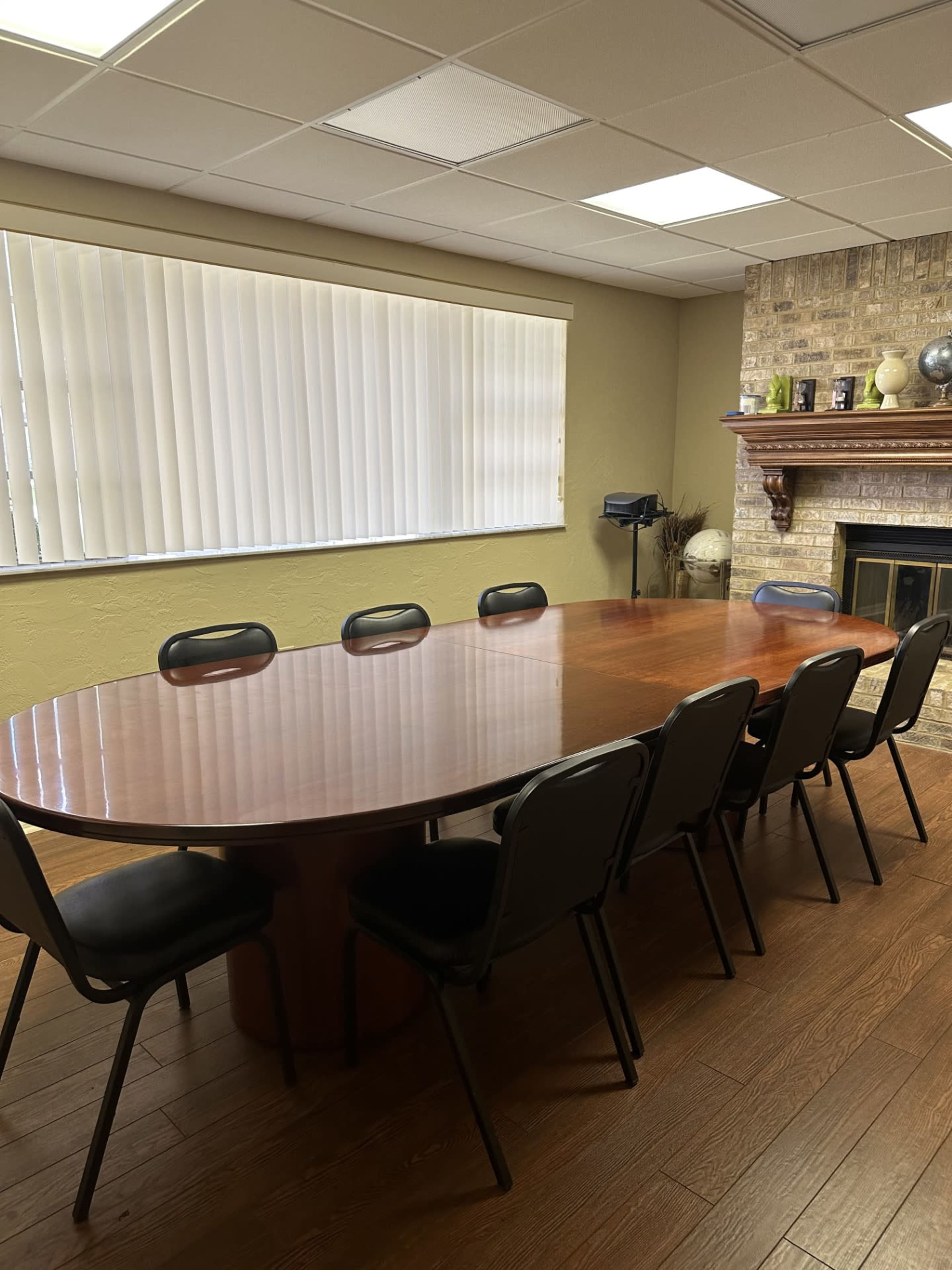 A large oval wooden conference table is surrounded by eight black chairs in a meeting room with a fireplace and window blinds.