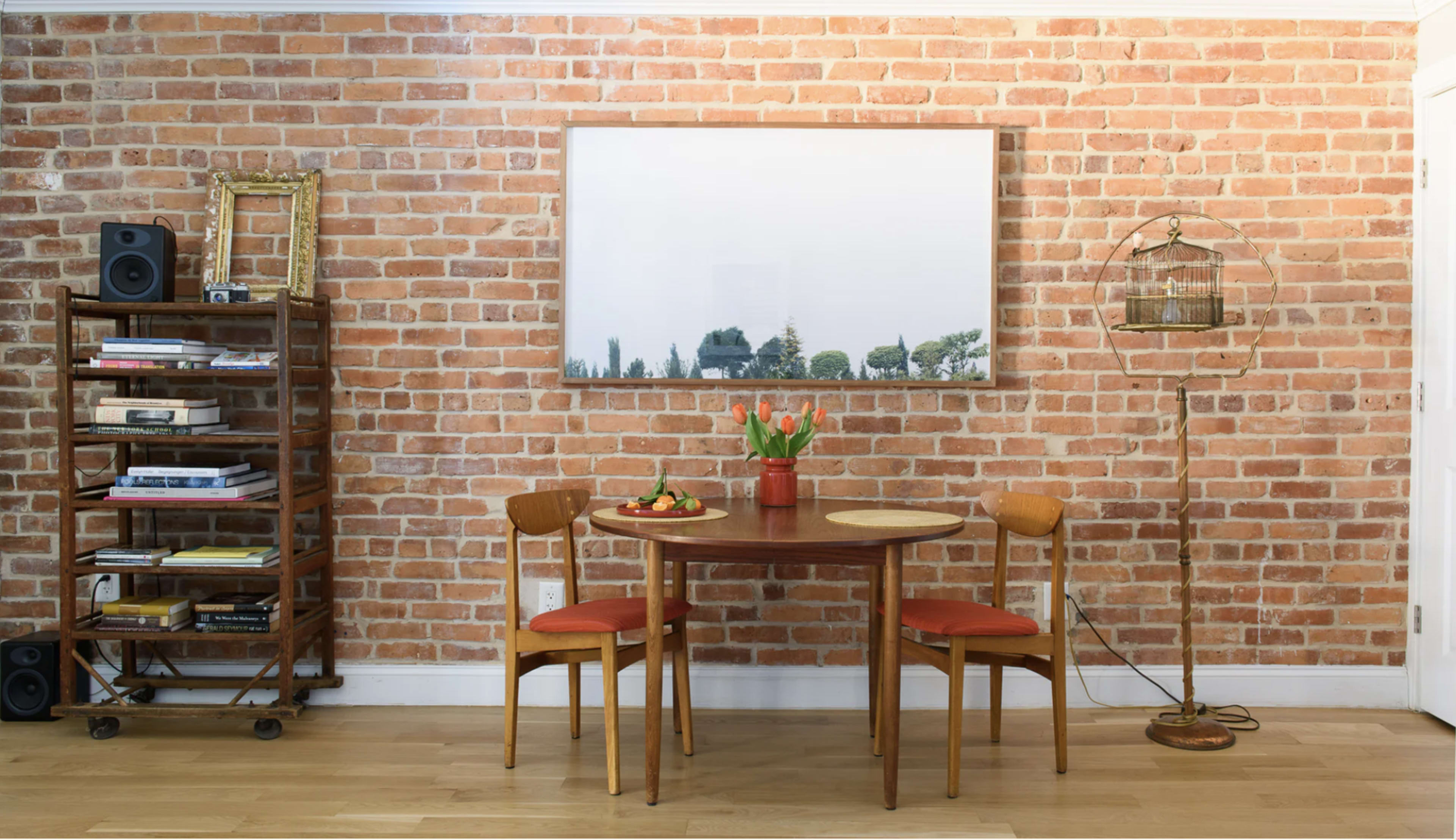 A wooden dining table with two chairs sits in front of a brick wall, flanked by a bookshelf and a decorative birdcage.