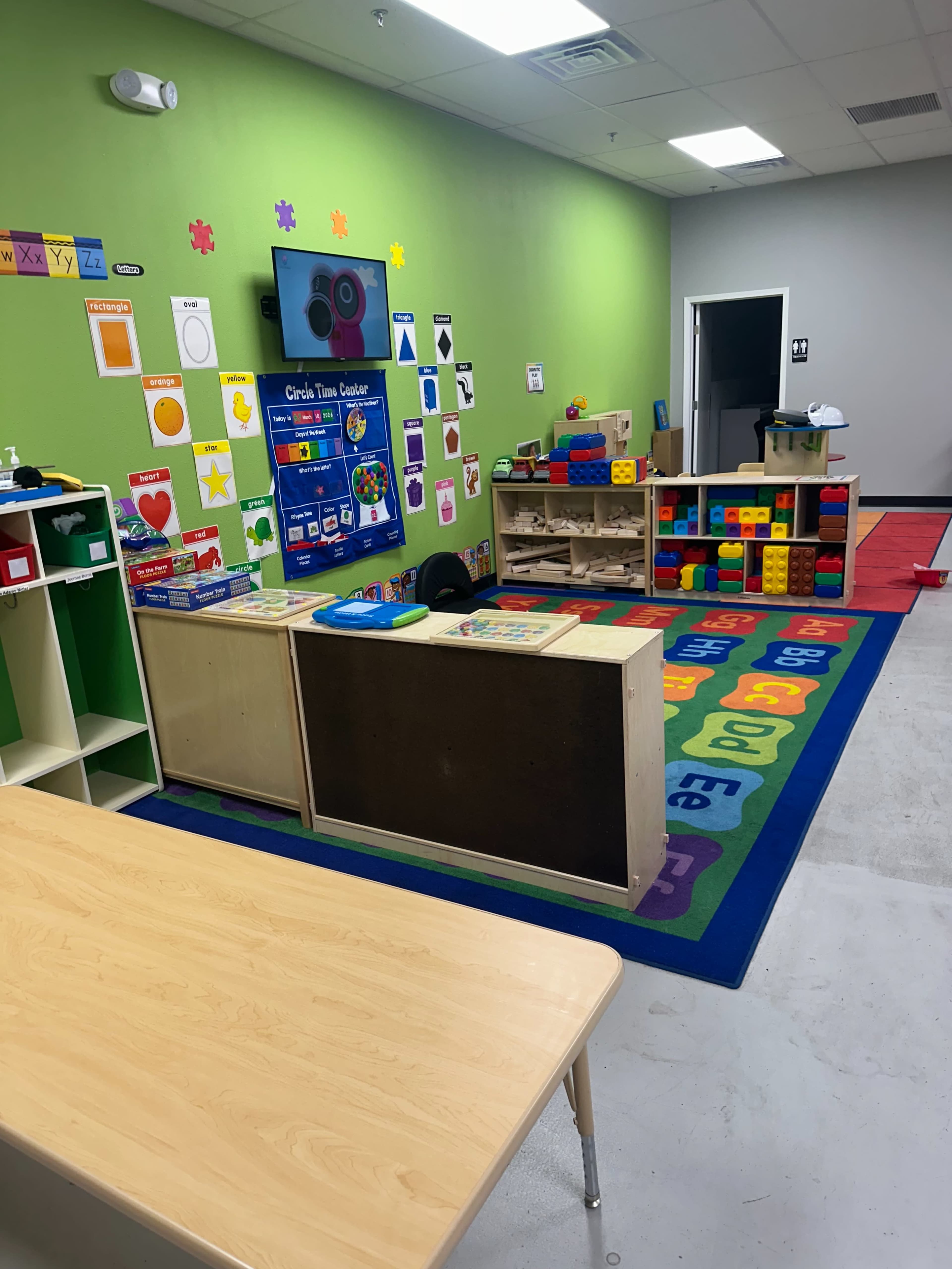 The image shows a brightly colored classroom with educational posters, a carpet with letters and numbers, and various toys and learning materials arranged on shelves.