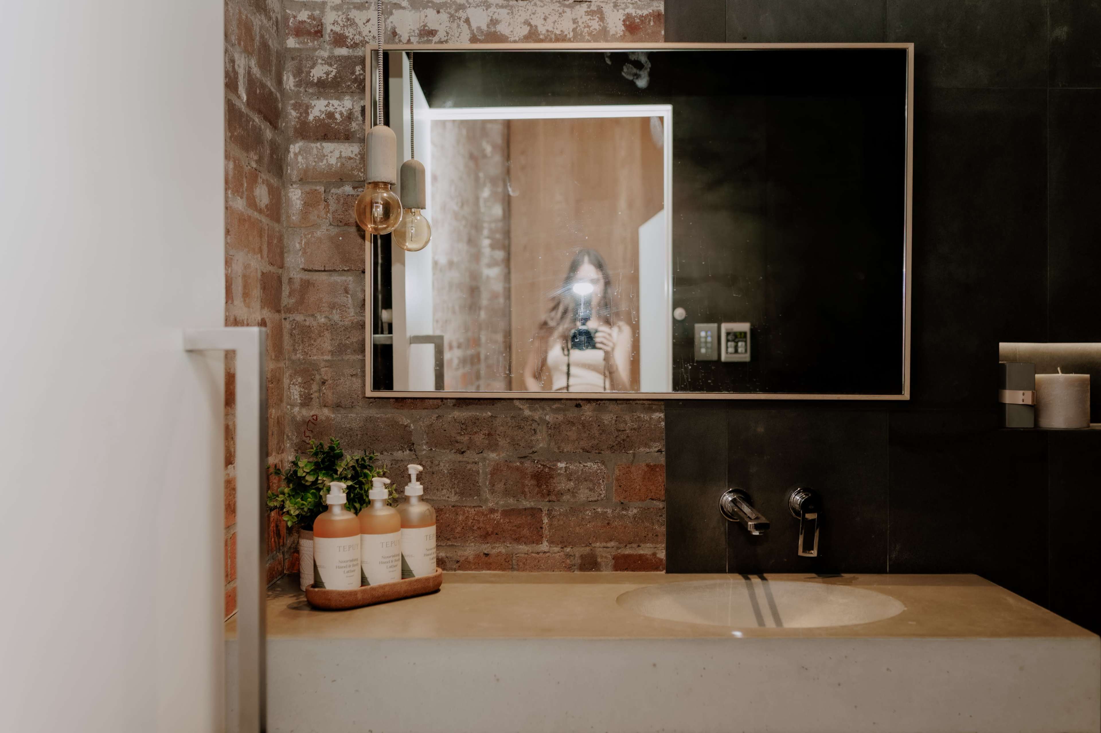 A modern bathroom features a concrete sink, a large mirror reflecting a person with a camera, and a wooden shelf with soap dispensers.