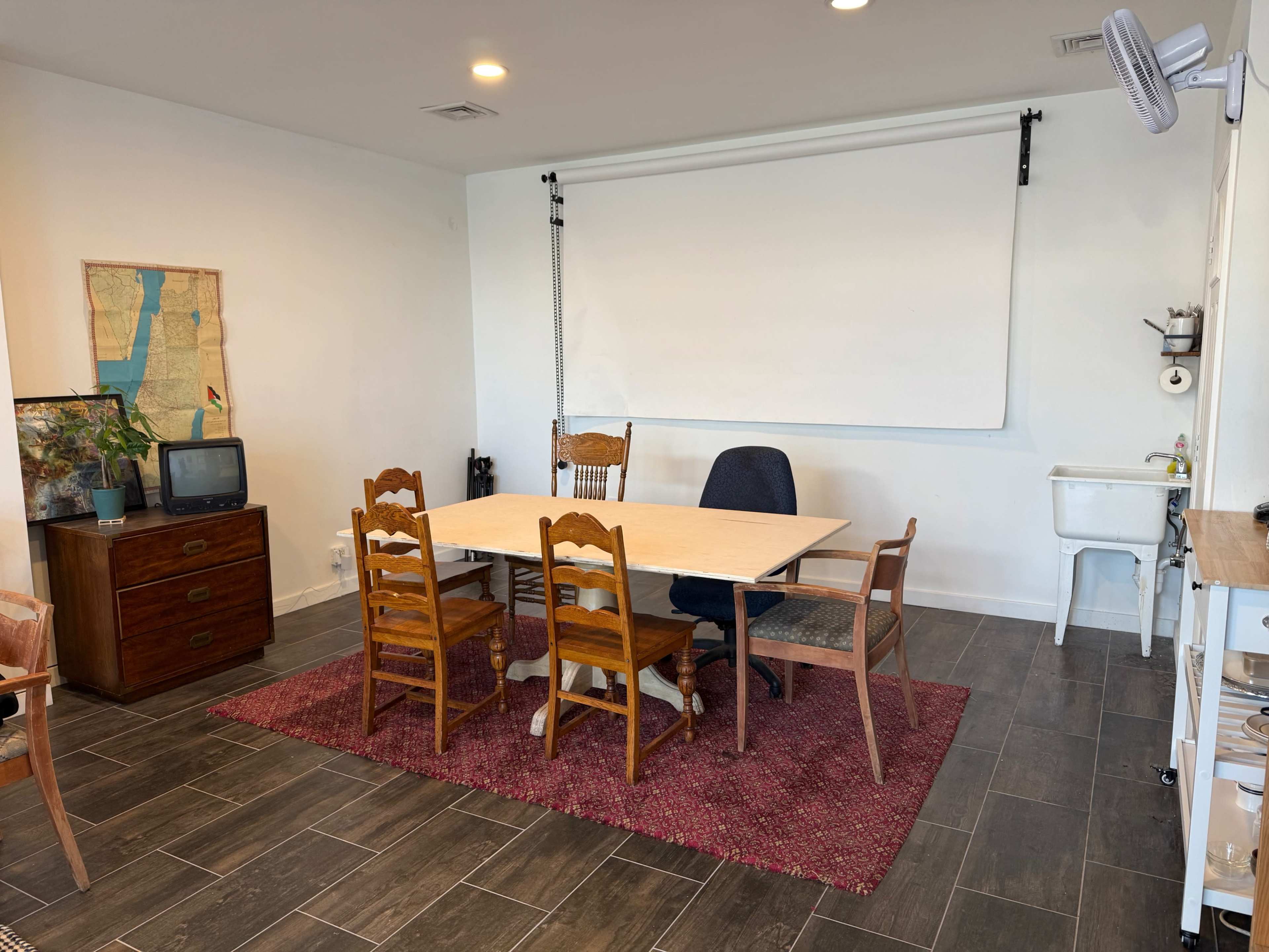 The image shows a brightly lit meeting room with a large table surrounded by several chairs, a small vintage television, and a wall-mounted projection screen.