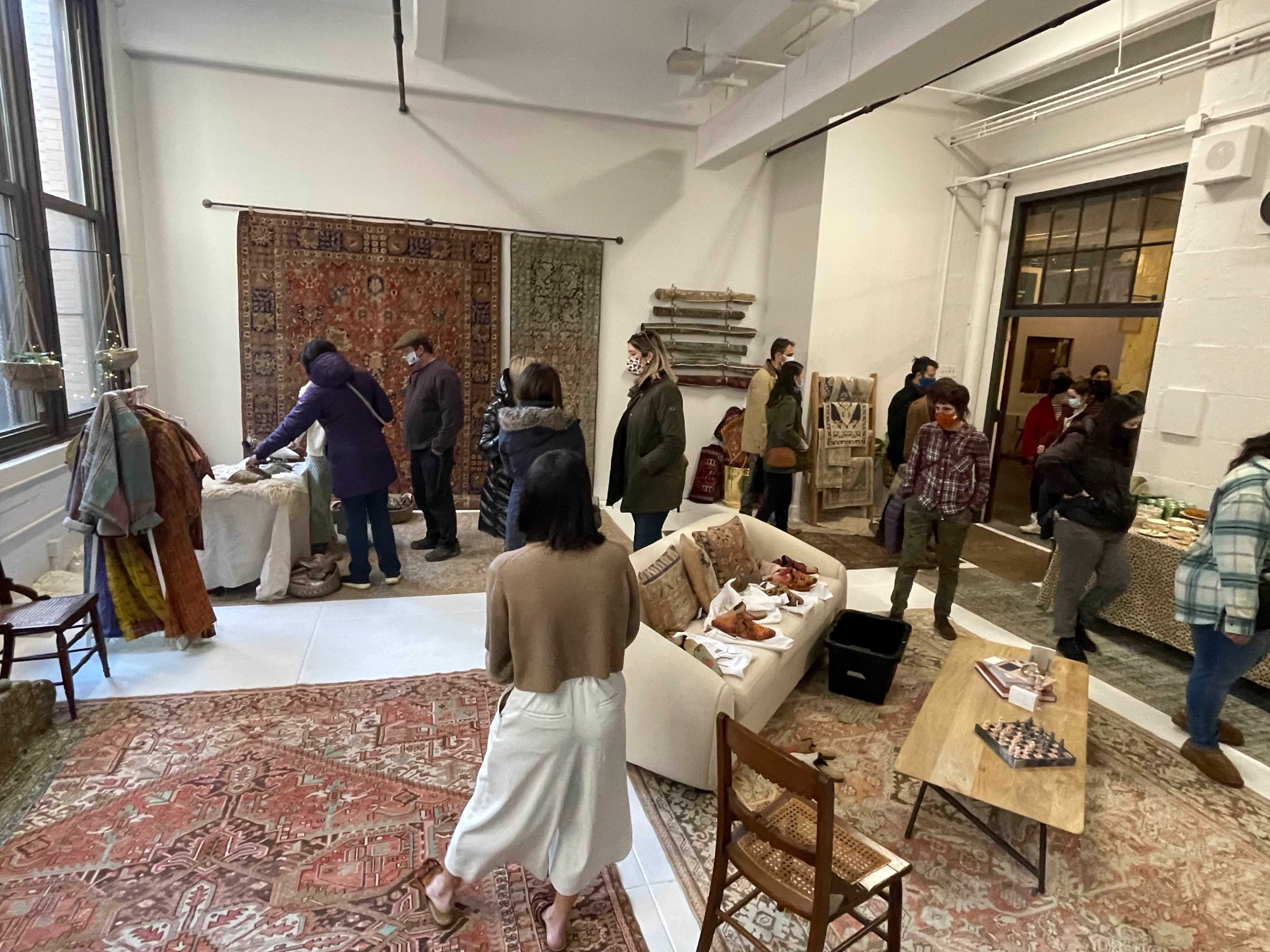 A group of people browse and interact in a spacious room decorated with rugs and textiles while displaying various items on tables.