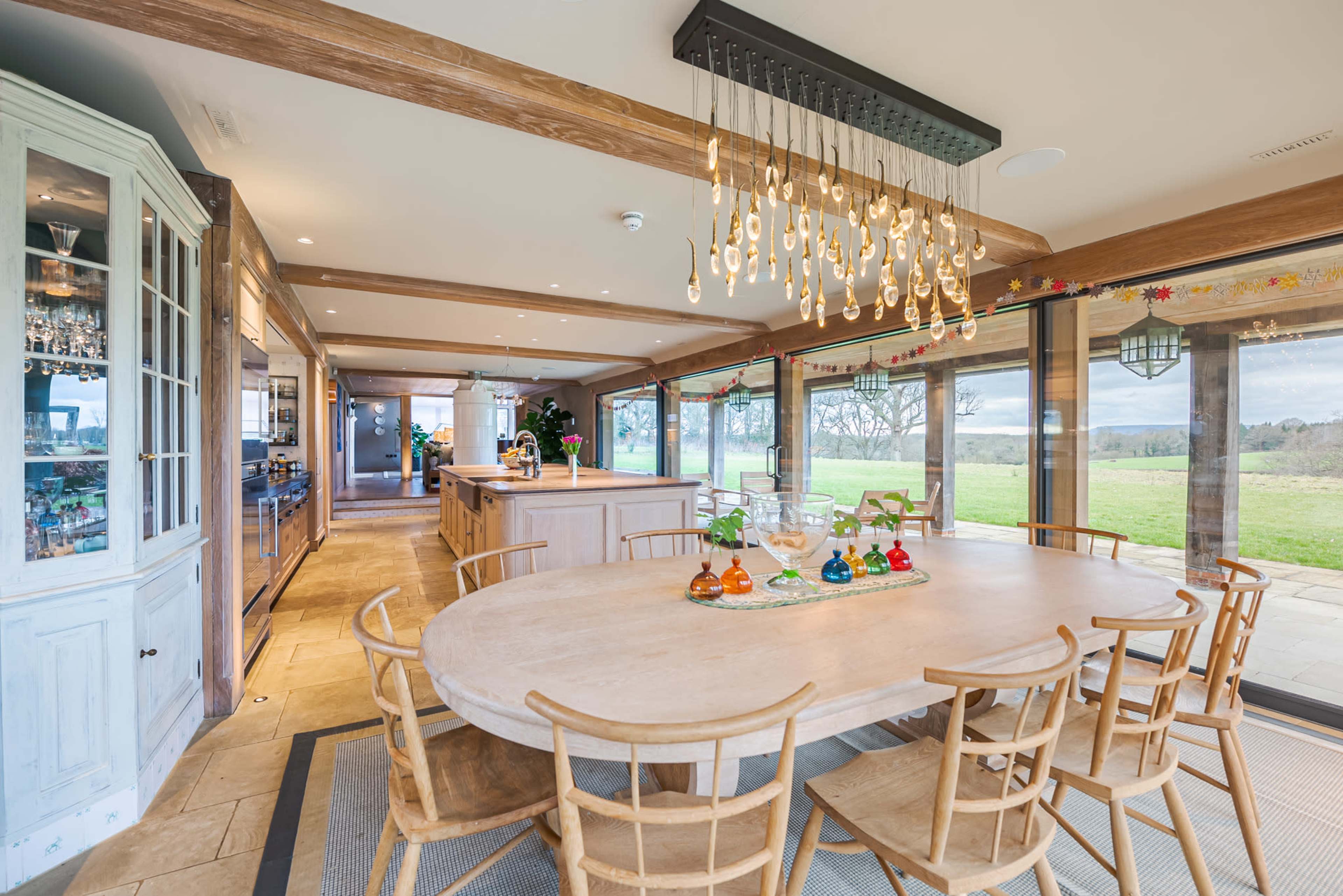 A spacious kitchen and dining area features a round wooden table surrounded by chairs, with large windows providing views of the outdoor landscape.