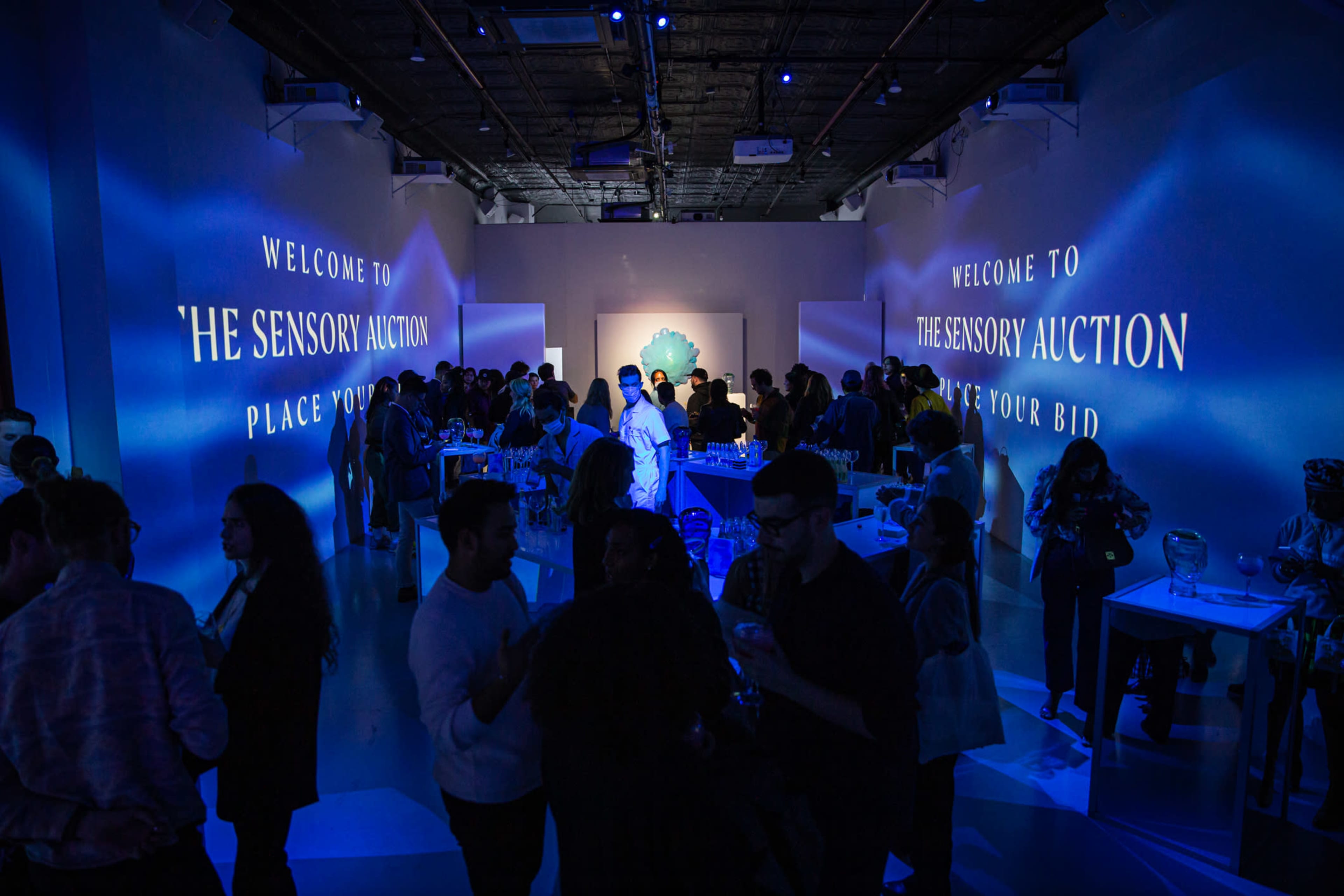 A crowd gathers in a brightly lit gallery space during an event titled "The Sensory Auction," characterized by blue lighting and large projections on the walls.