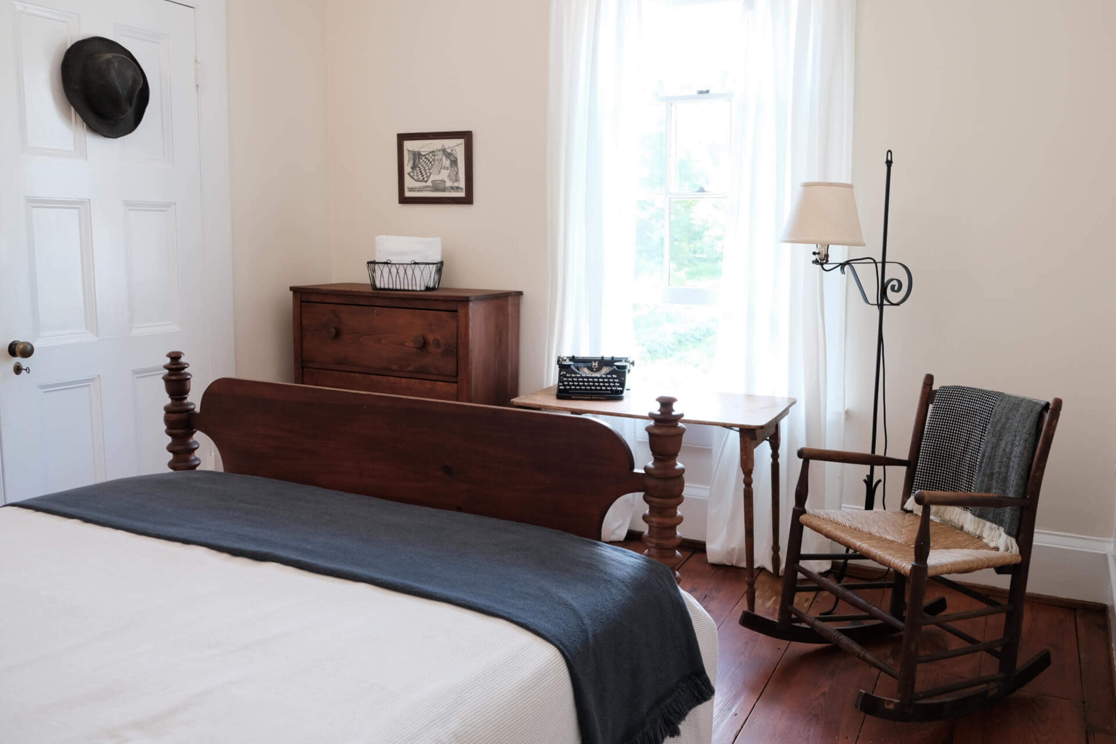 A simple bedroom featuring a wooden bed, a rocking chair, a dresser, and a small table with a typewriter, all illuminated by natural light from a window.
