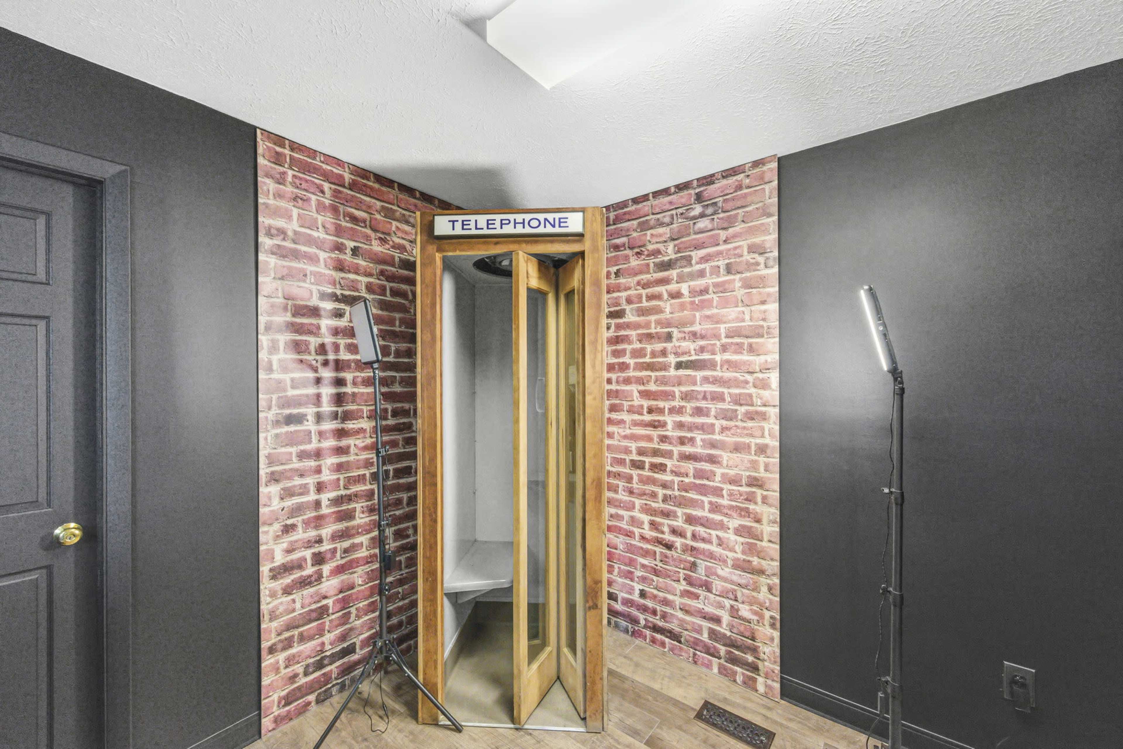 The image shows a vintage telephone booth made of wood, situated in a corner against a textured brick wall.