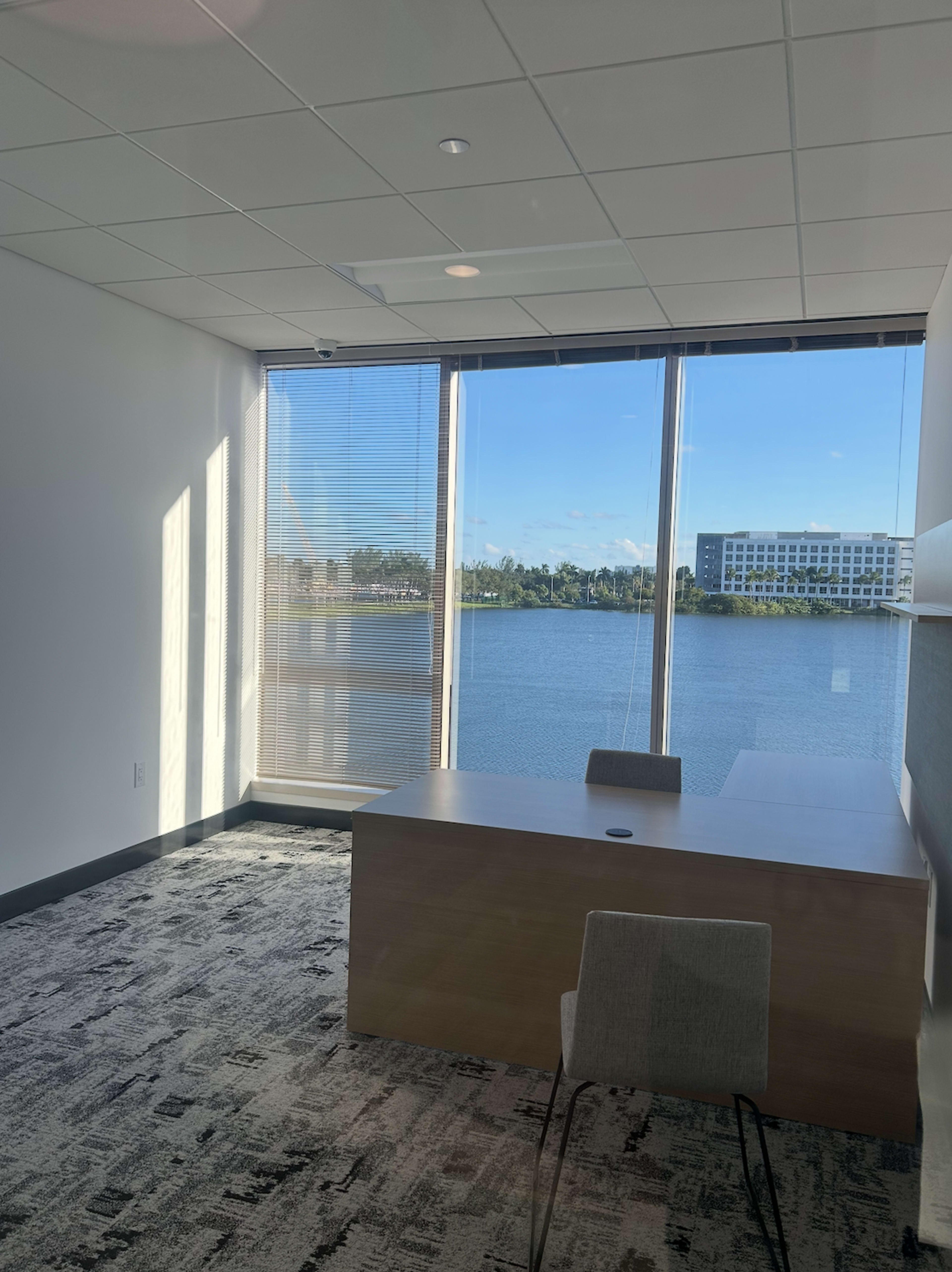 The image shows a modern office space with a wooden desk and a chair, facing large windows that overlook a body of water and foliage outside.
