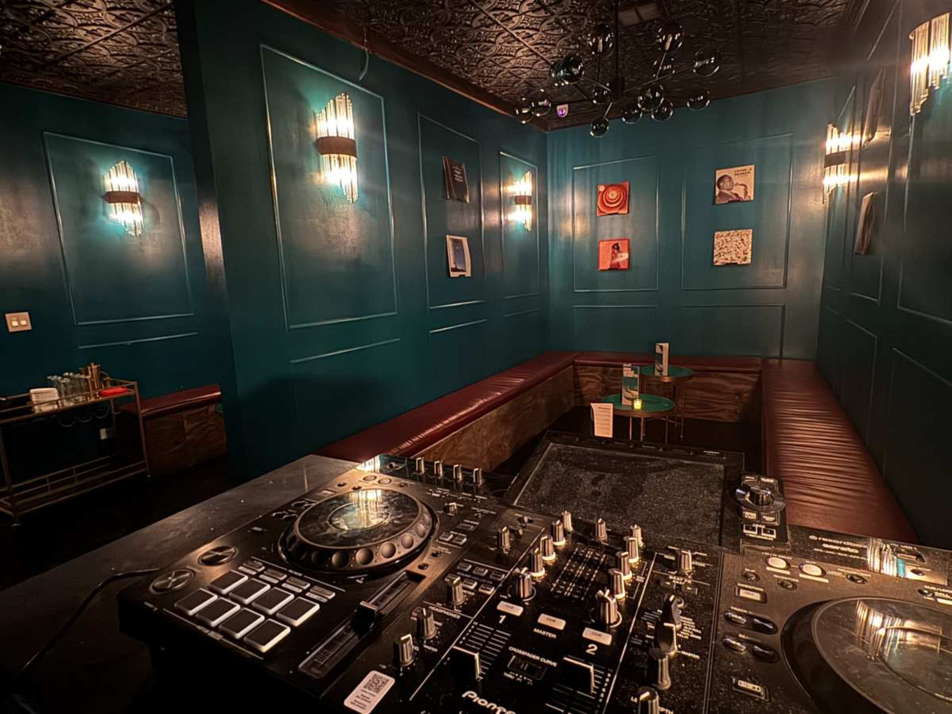 The image shows a dimly lit room with teal walls, a DJ setup featuring turntables and mixers, and leather seating along the walls.
