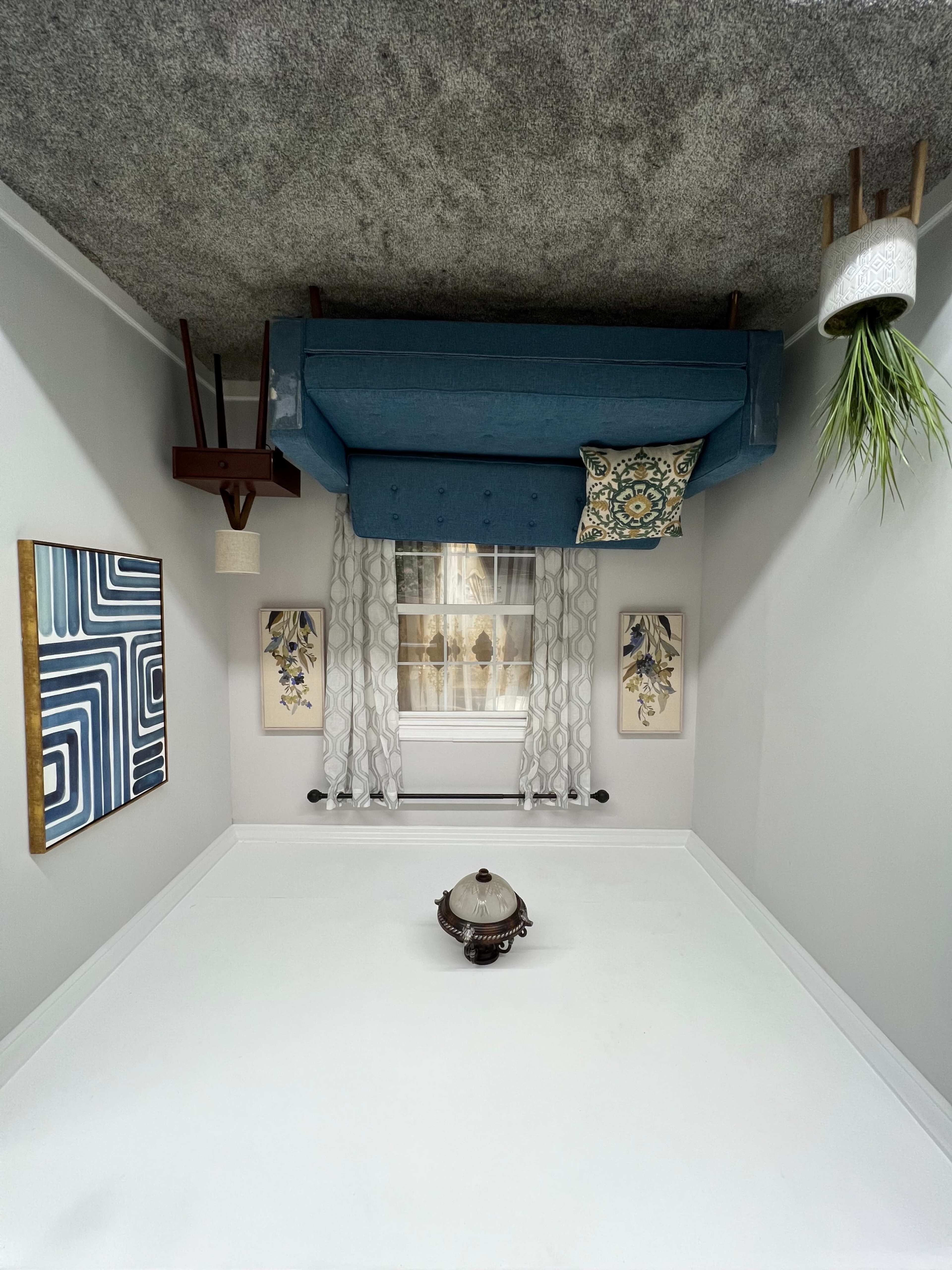 An inverted room with a blue couch mounted on the ceiling, accompanied by decorative wall art and a potted plant.
