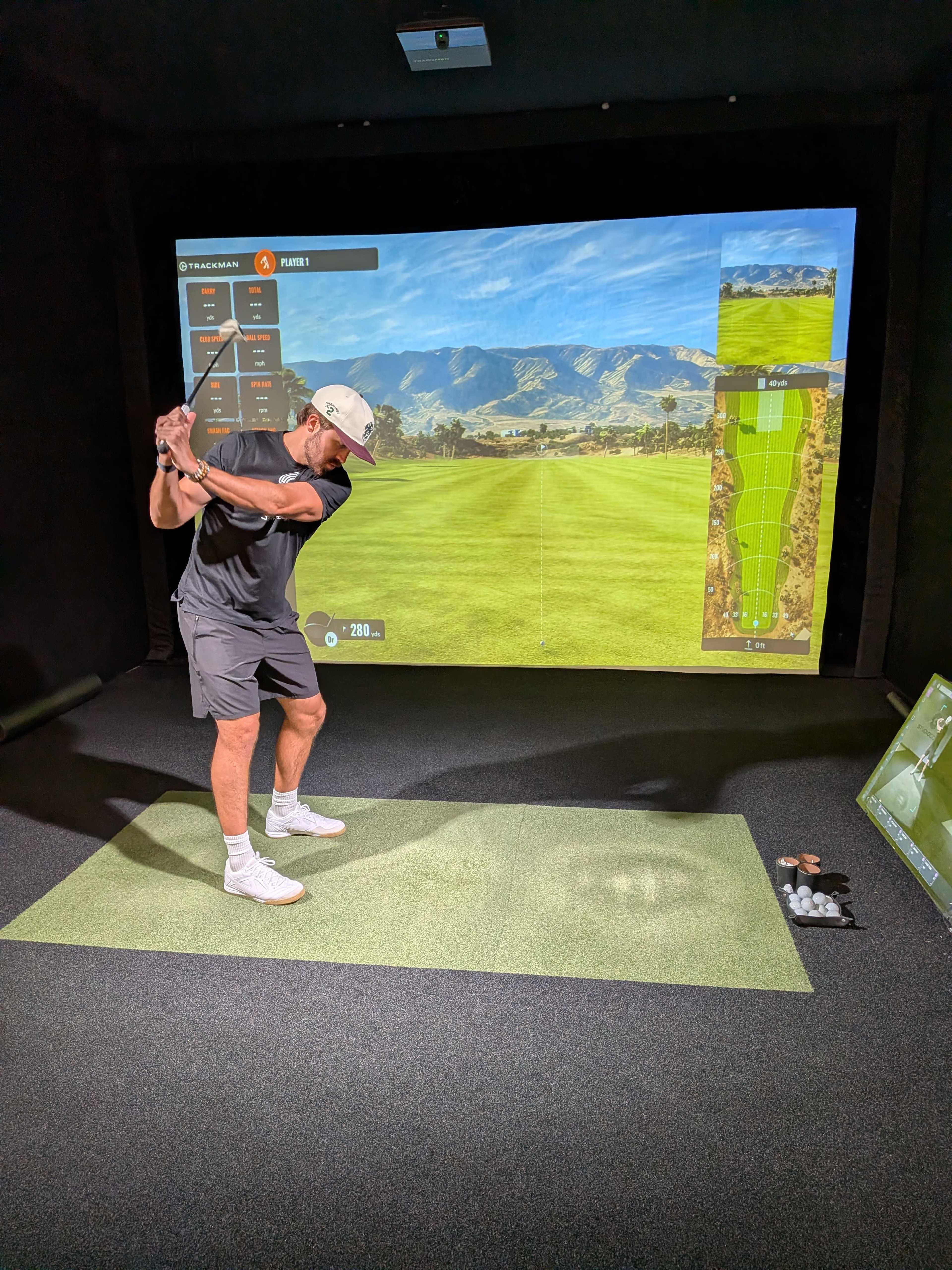 A golfer is taking a swing in an indoor simulation environment with a large screen displaying a golf course landscape.