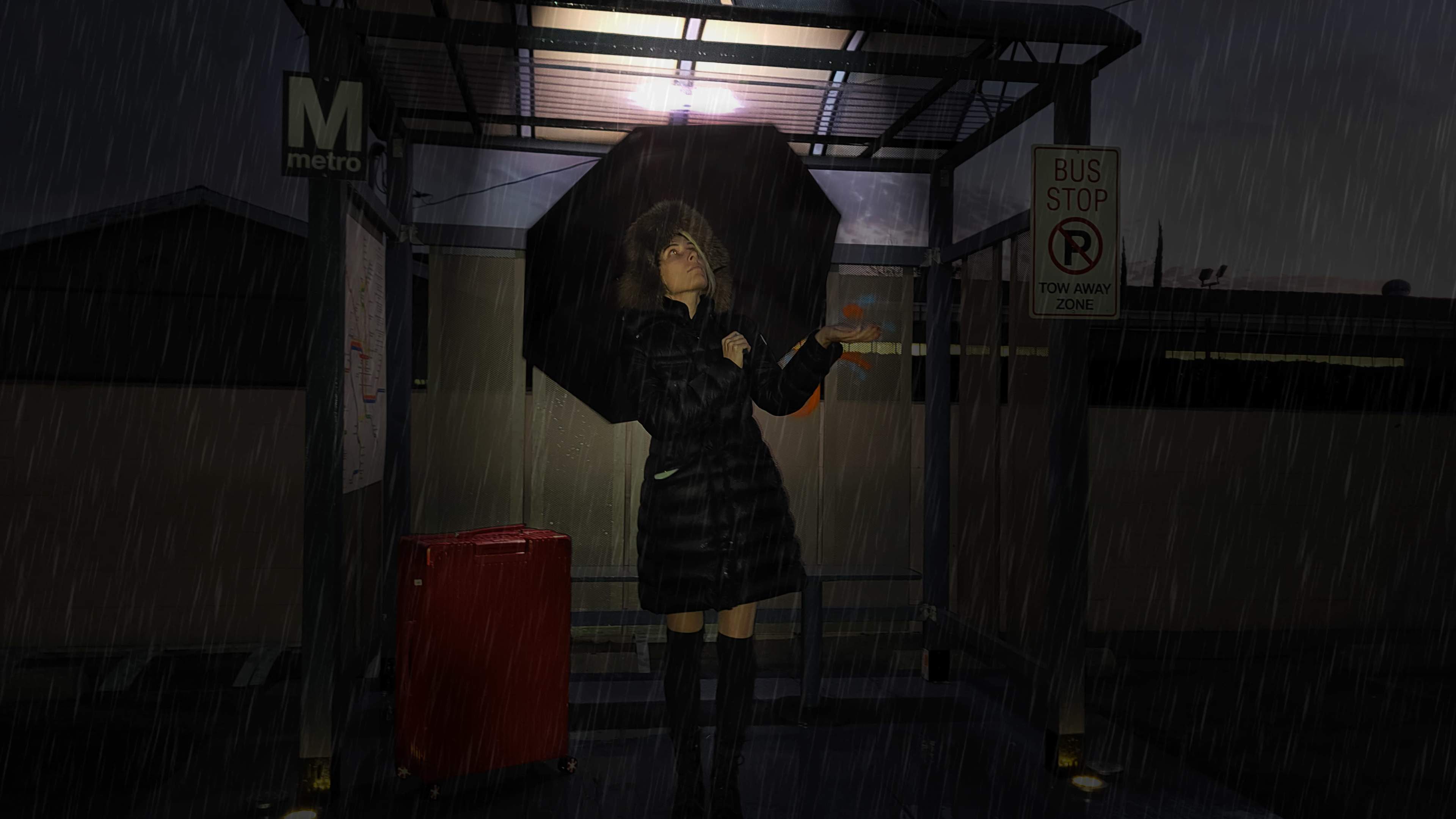 A woman holding an umbrella stands at a bus stop under a shelter while it rains, with a red suitcase next to her.