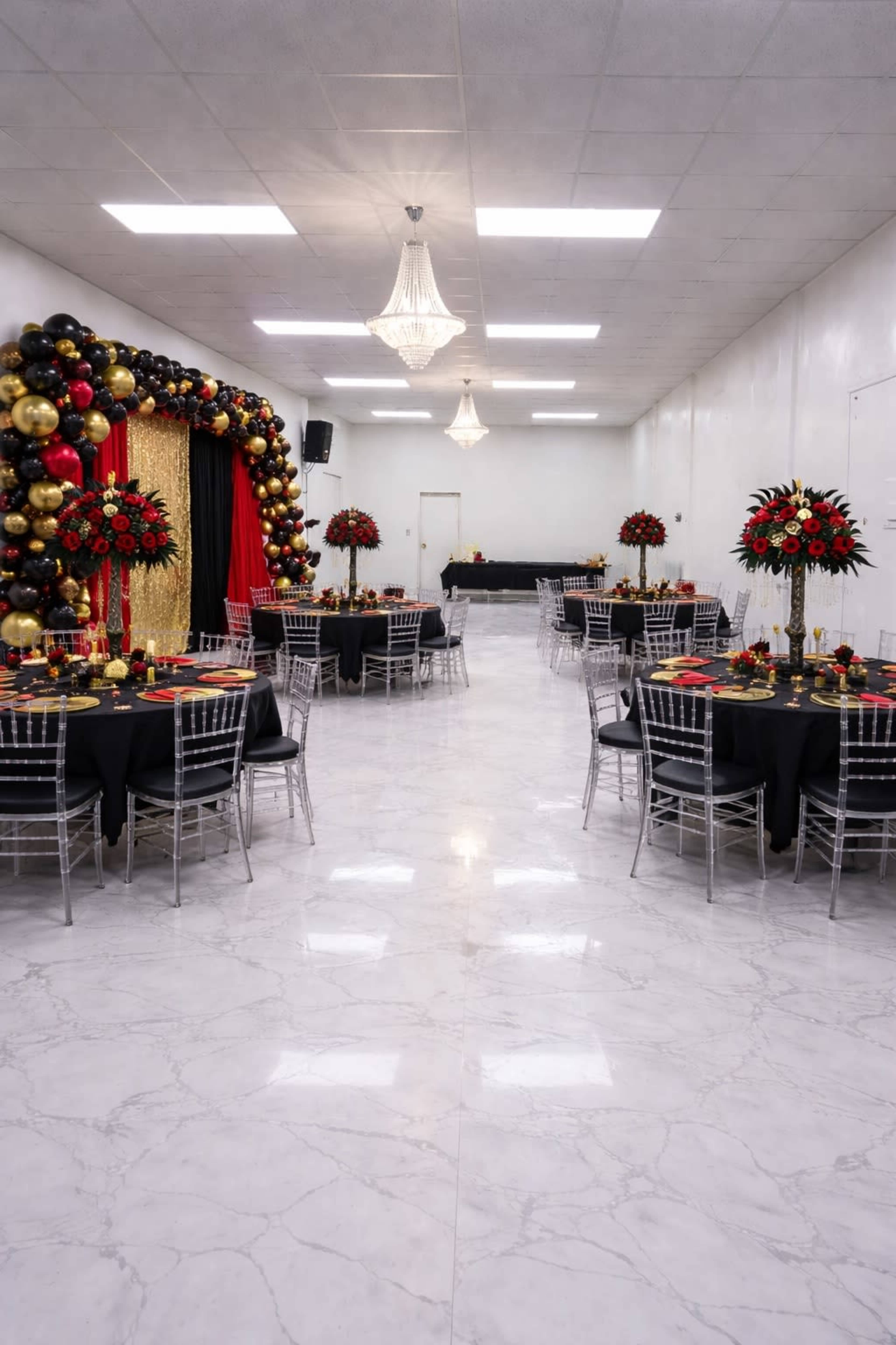 The image shows a banquet hall set up with elegant tables, each adorned with black tablecloths and tall floral centerpieces, along with a backdrop of black, gold, and red balloons.