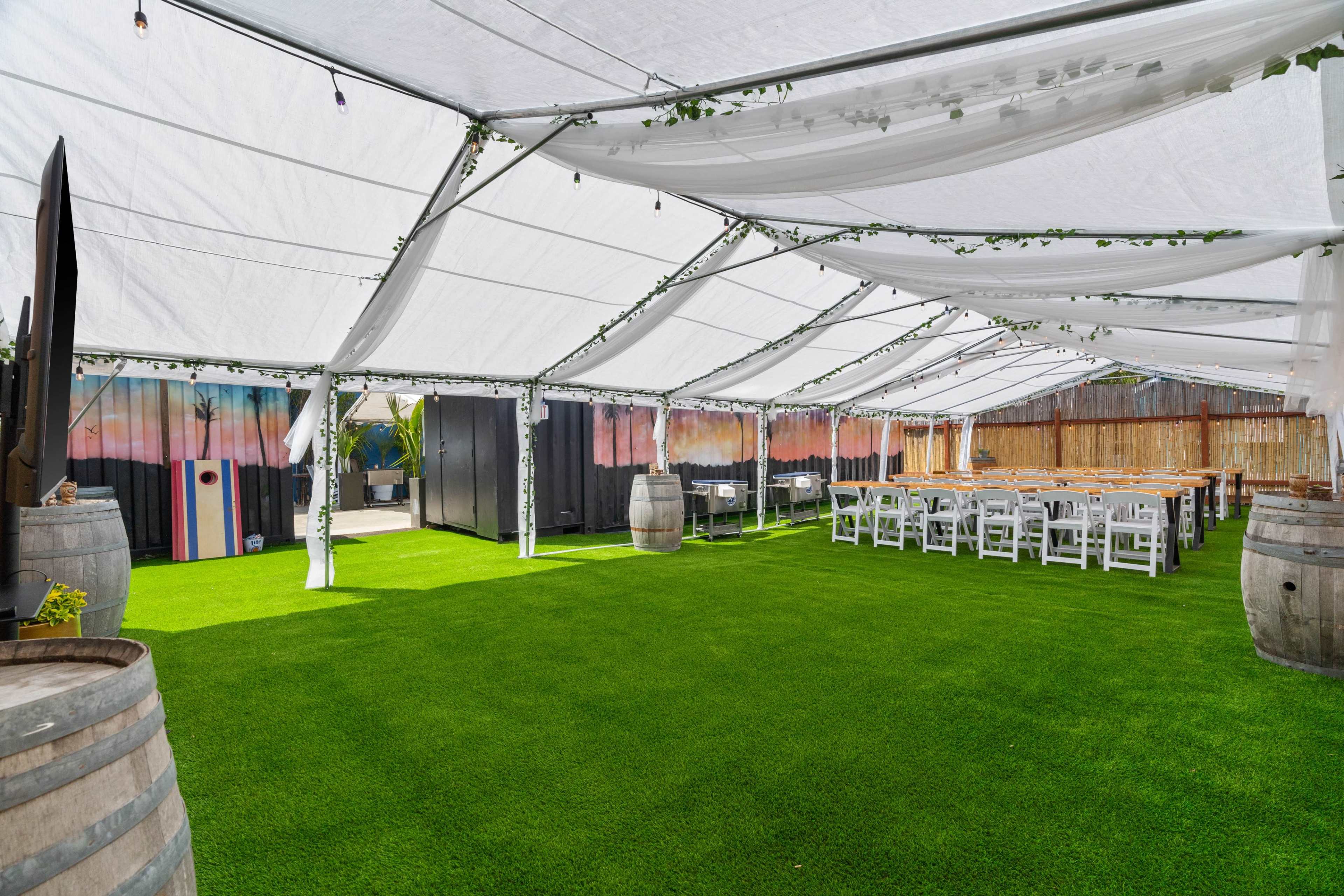 A large white tent with a green carpet covers tables and chairs set for an event, surrounded by wooden barrels.