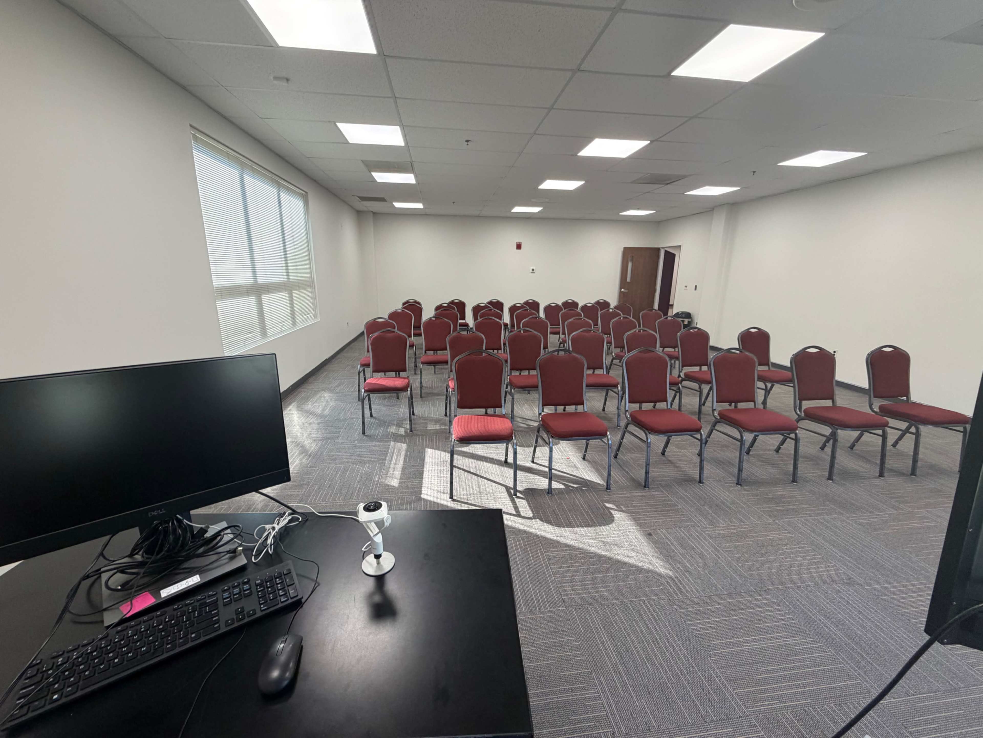 A conference room features rows of red chairs set up for an audience, with a desk and computer in the foreground.