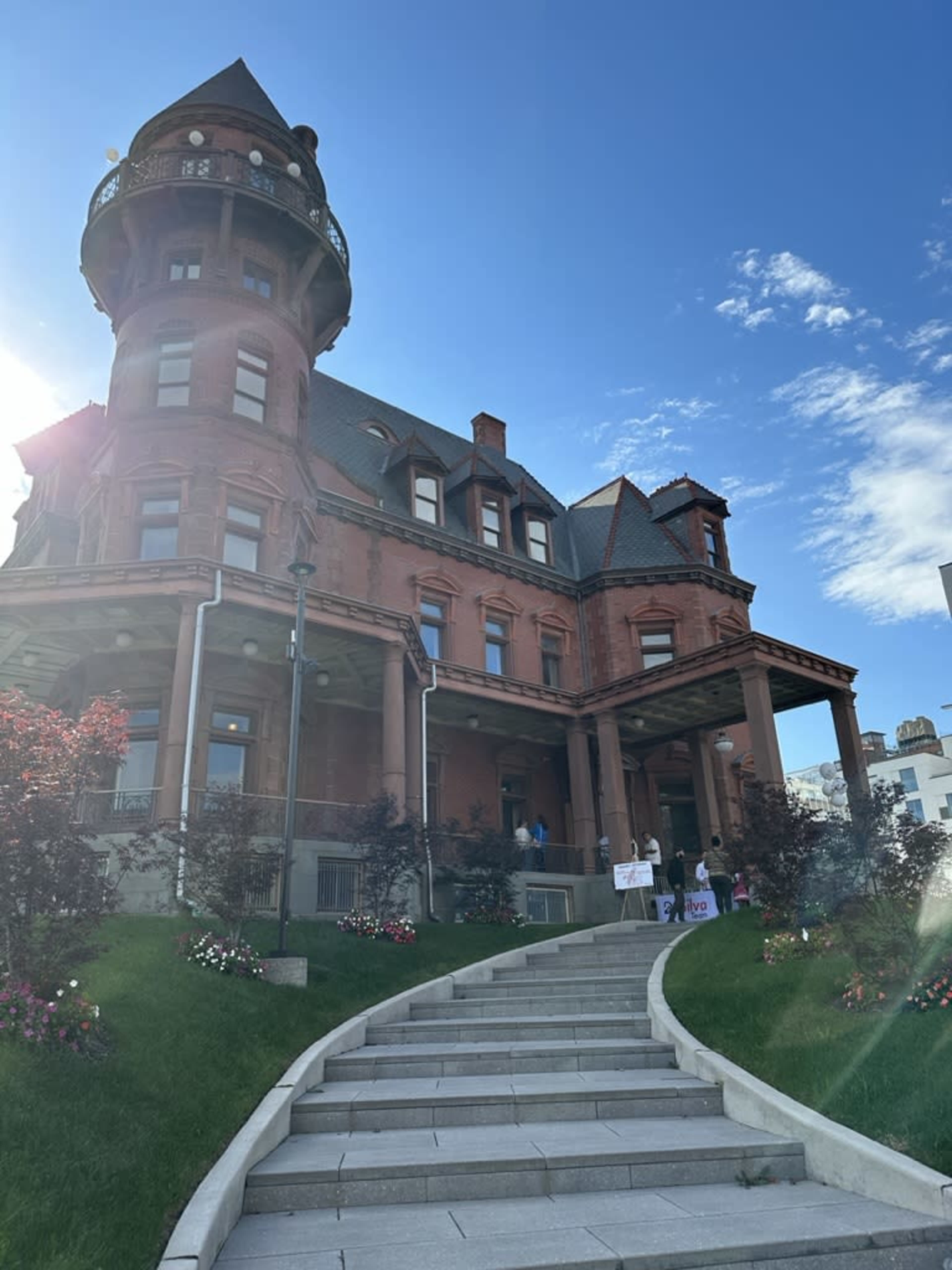 A large red brick mansion with a turret stands on a landscaped hillside, featuring a set of stairs leading to its entrance.