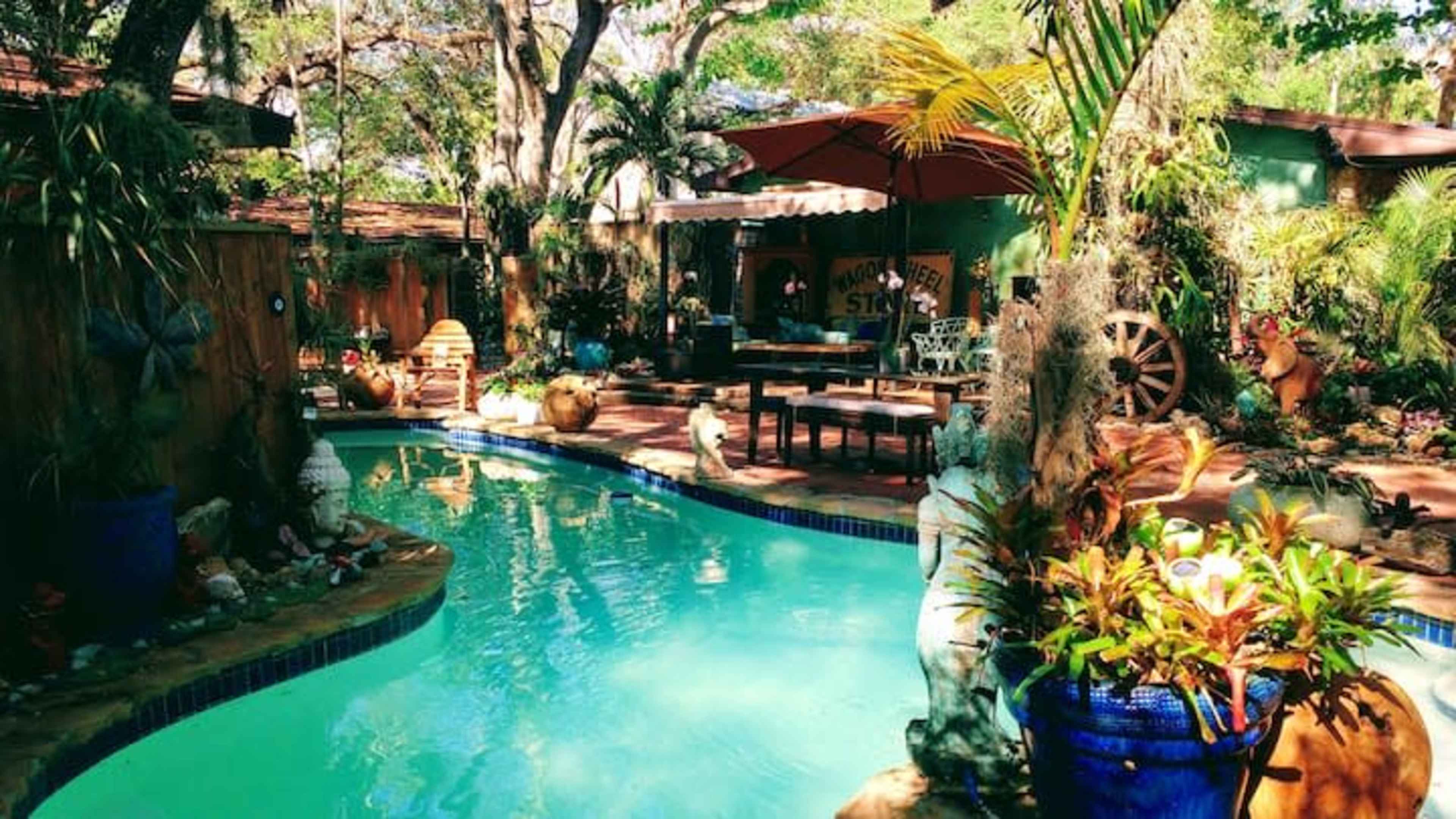 The image shows a tranquil backyard pool area surrounded by greenery, with a patio featuring outdoor furniture and decorations.